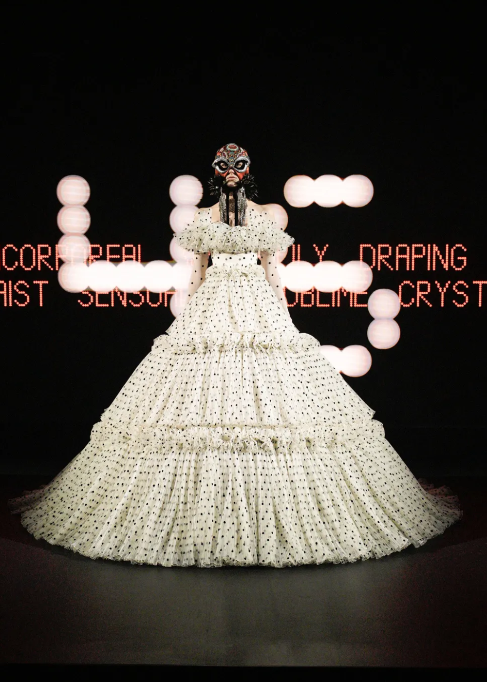 Modelo em vestido de bolinhas, máscara elaborada. Fundo com palavras em luzes. Semana de Alta-Costura 2025.