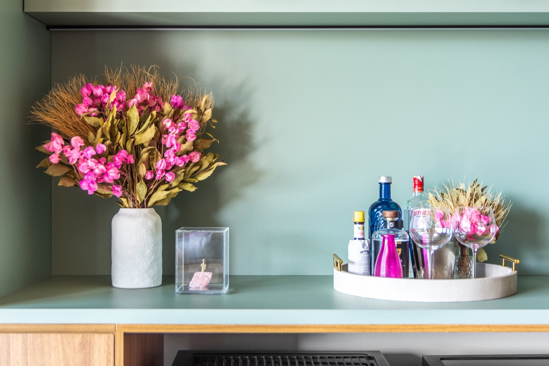 Flores cor-de-rosa em vaso, garrafas e copos decoram balcão verde na decoração.