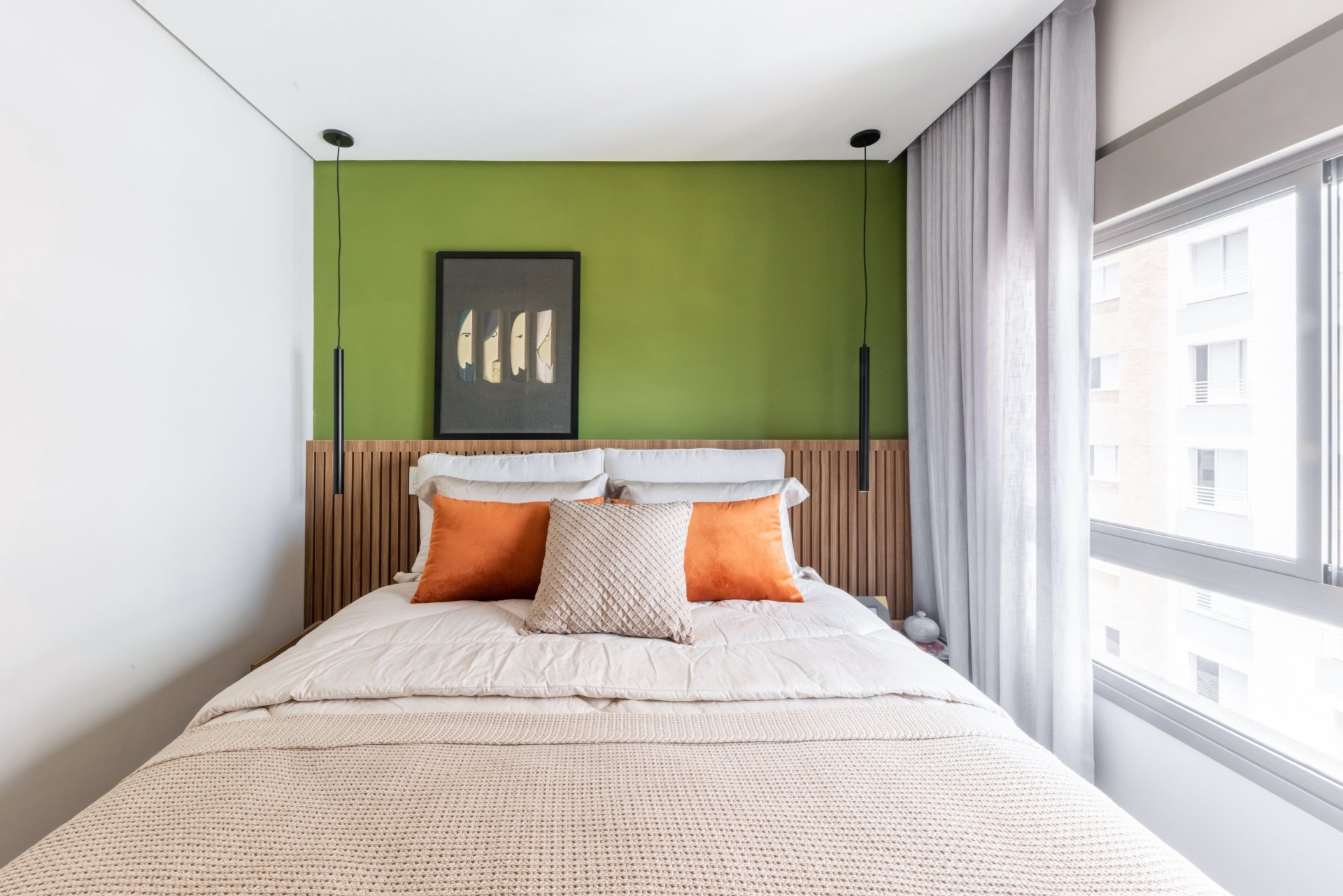 Quarto com parede verde, cabeceira de ripas e cama com almofadas laranja e bege. Janela ao lado direito com cortina cinza. Verde na decoração