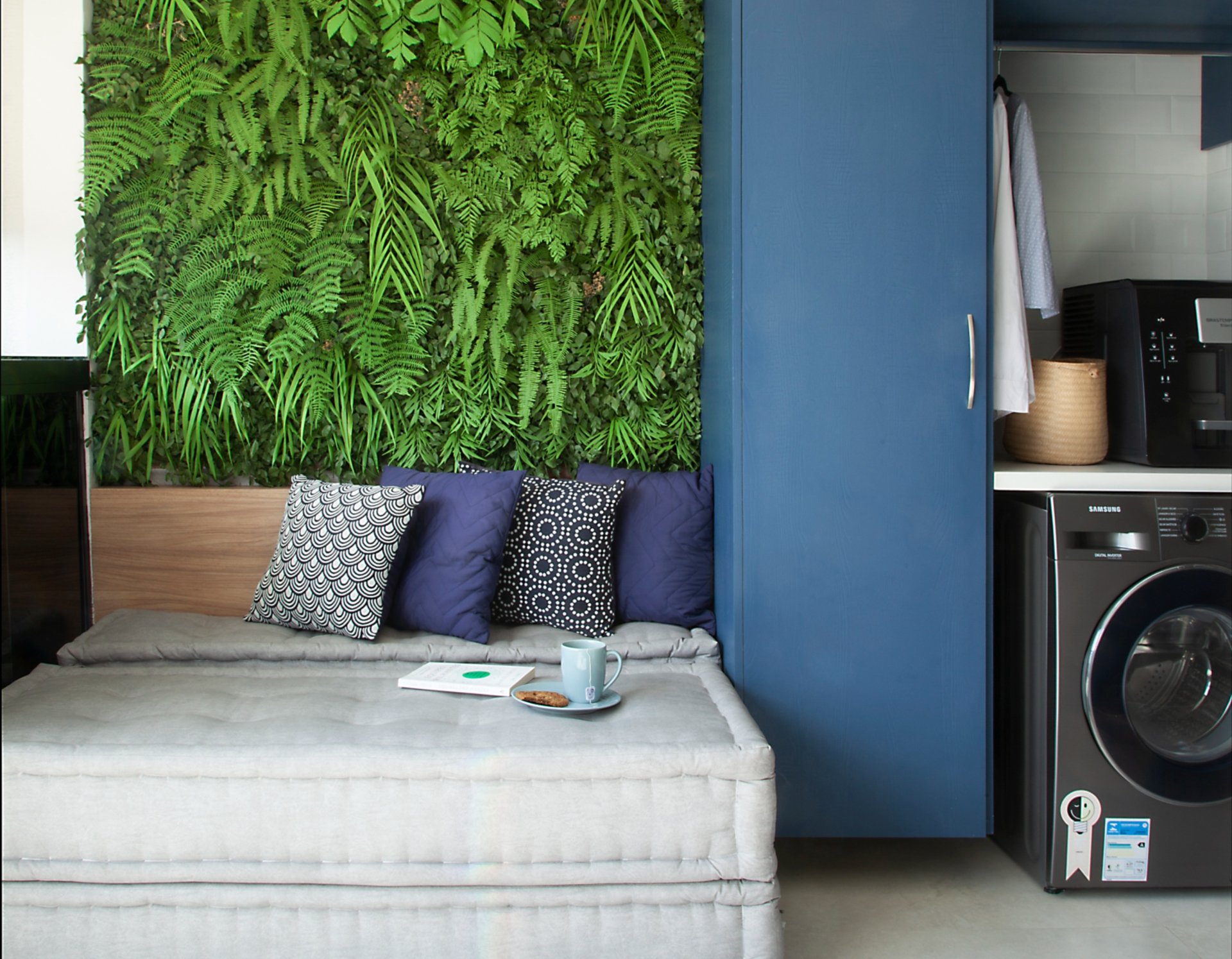 Sofá com almofadas em frente a parede verde de plantas. Armário azul ao lado, máquina de lavar na direita. Verde na decoração