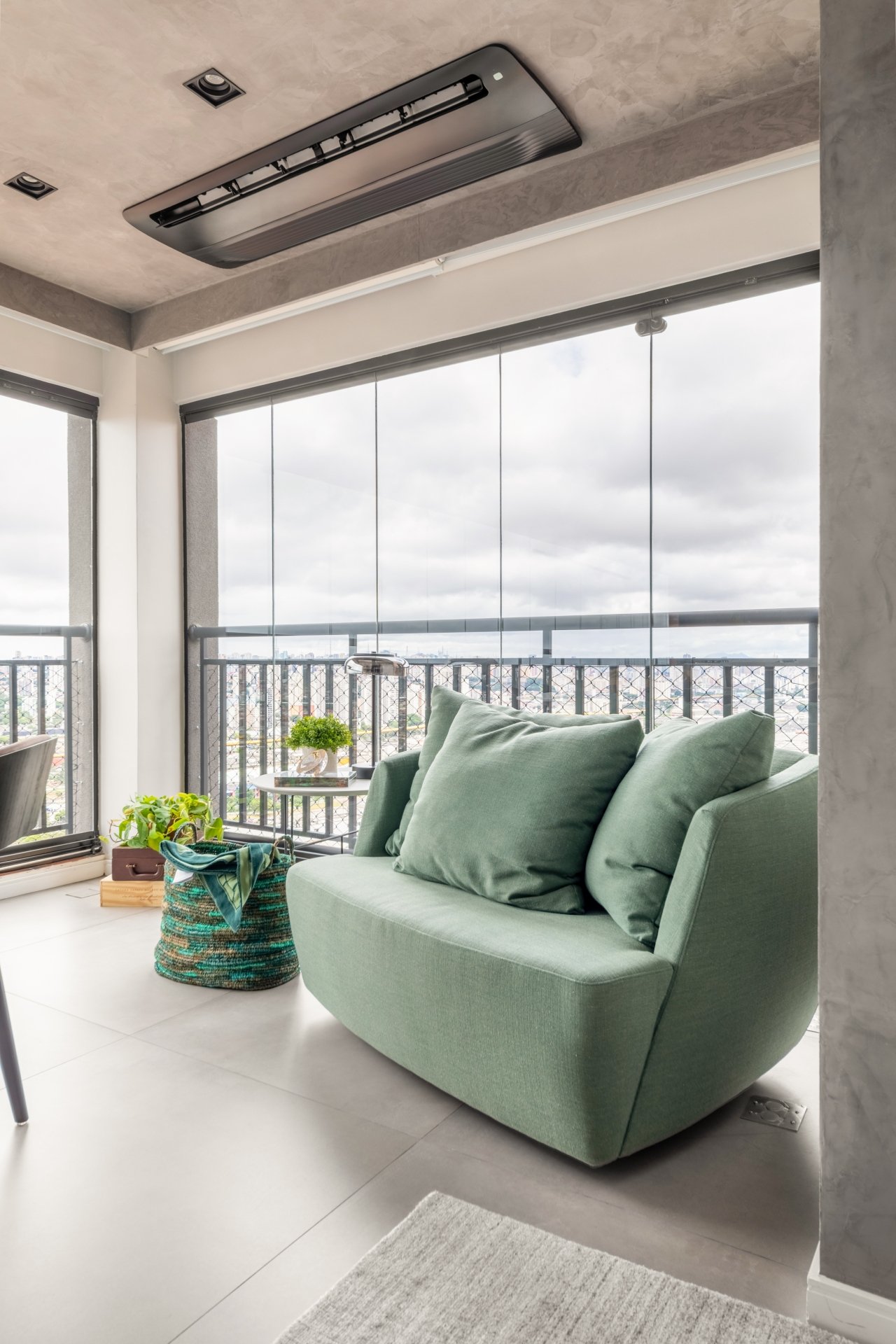 Sofá verde em varanda com janelas de vidro, plantas decorativas e cesta tricotada verde na decoração ao lado.