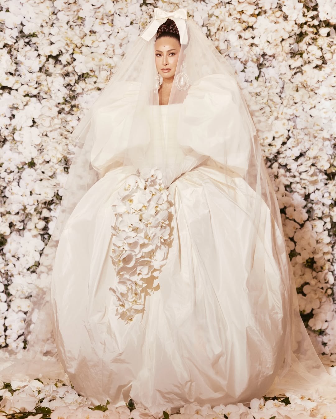 Vestido de noiva volumoso com detalhes florais e véu, em um fundo de flores brancas.