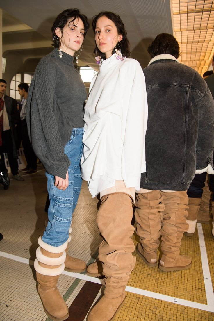 Duas modelos usando roupas da Y/Project, com suéter, jeans e botas longas em ambiente interno.