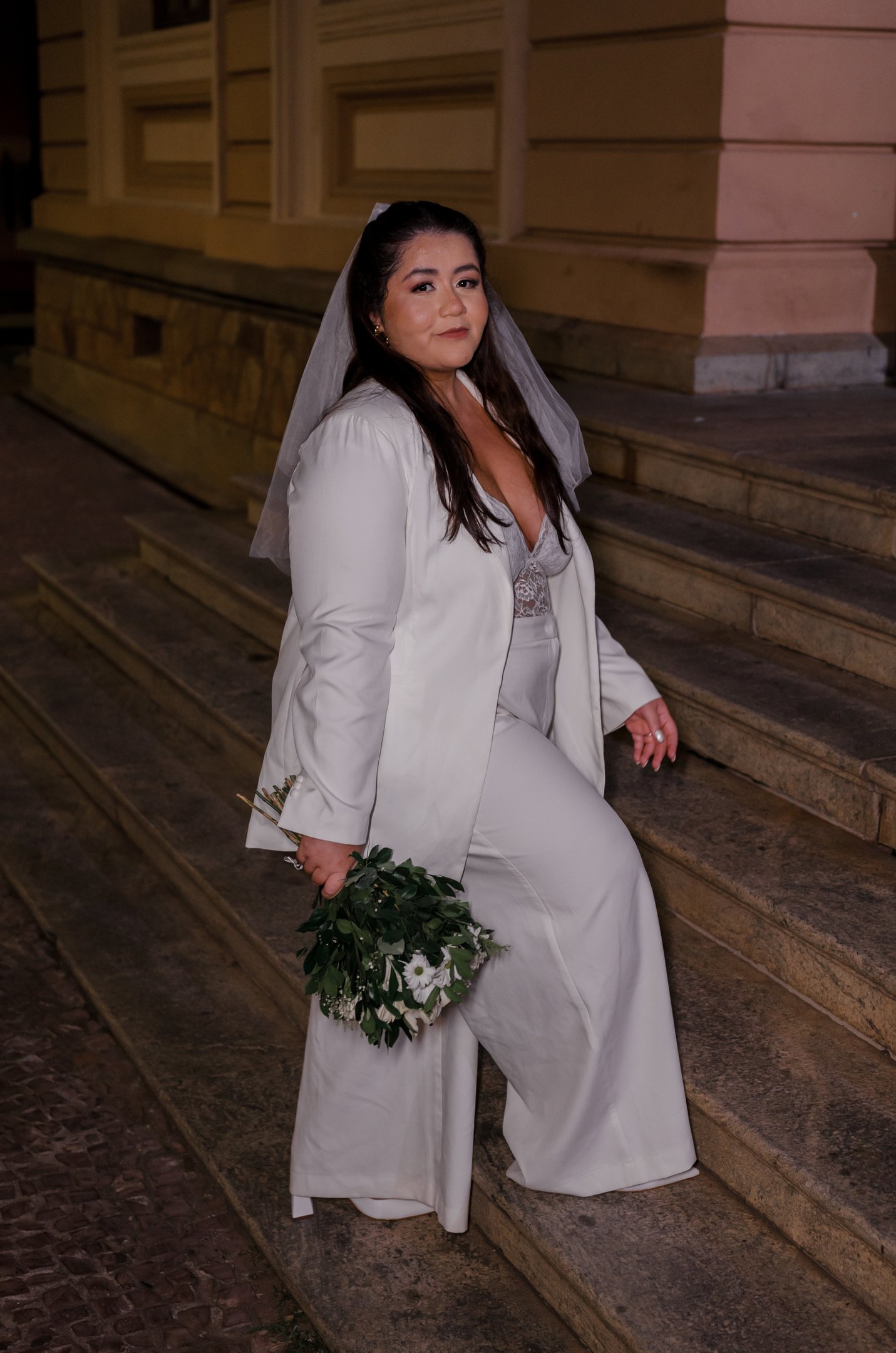 Looks de noiva plus size: layla posa em escada com buquê, usando terno branco e véu.