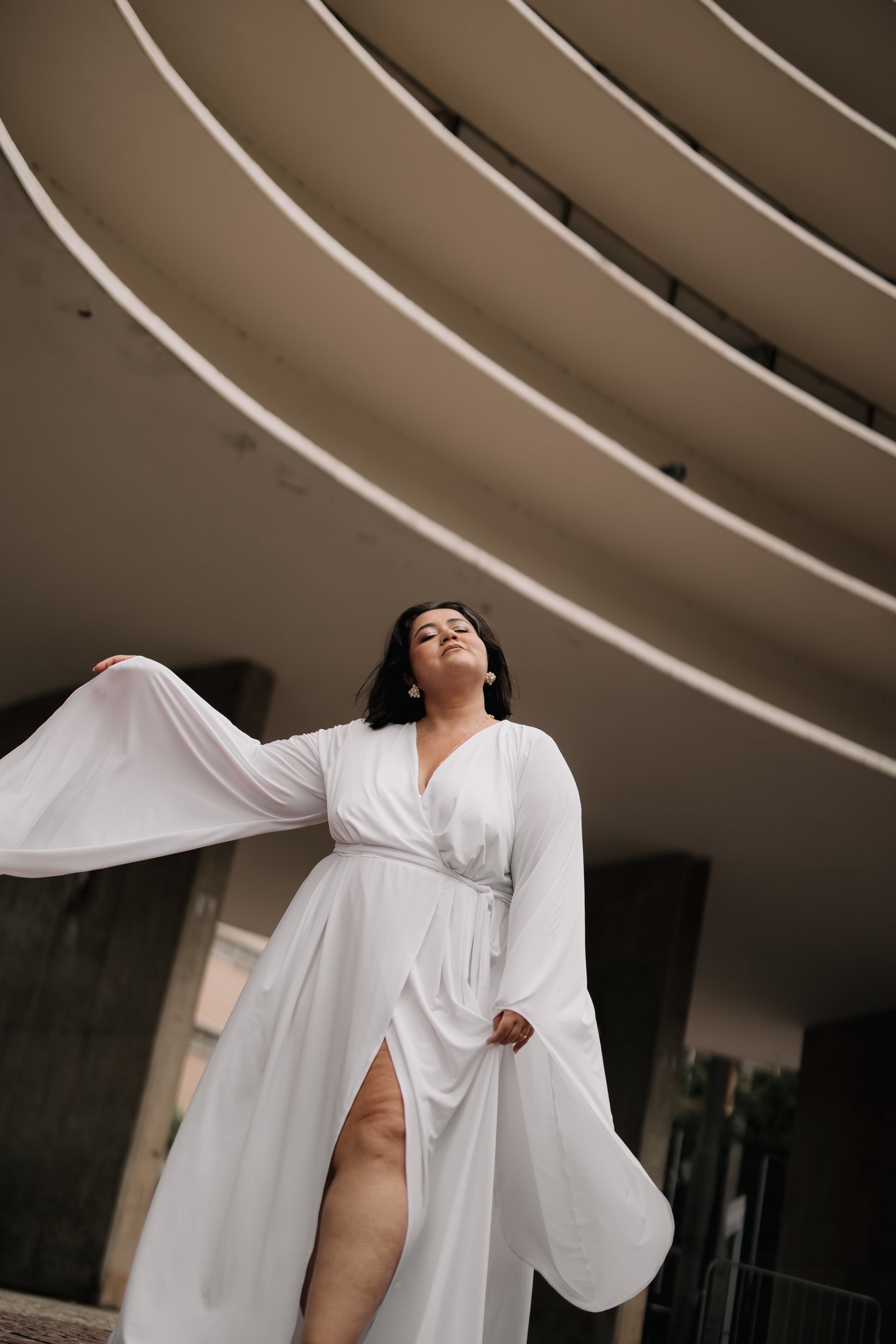 Vestido branco elegante com mangas longas em frente a uma estrutura arquitetônica moderna, ideal para looks de noiva plus size.