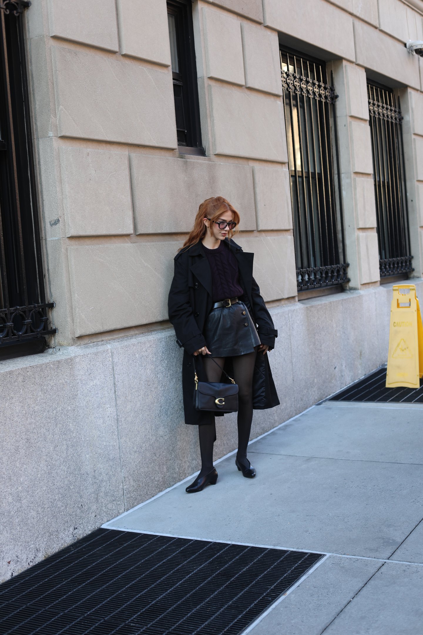 Jordanna Maia usando casaco longo preto, saia de couro, botas e bolsa preta, destacando tendência do inverno.