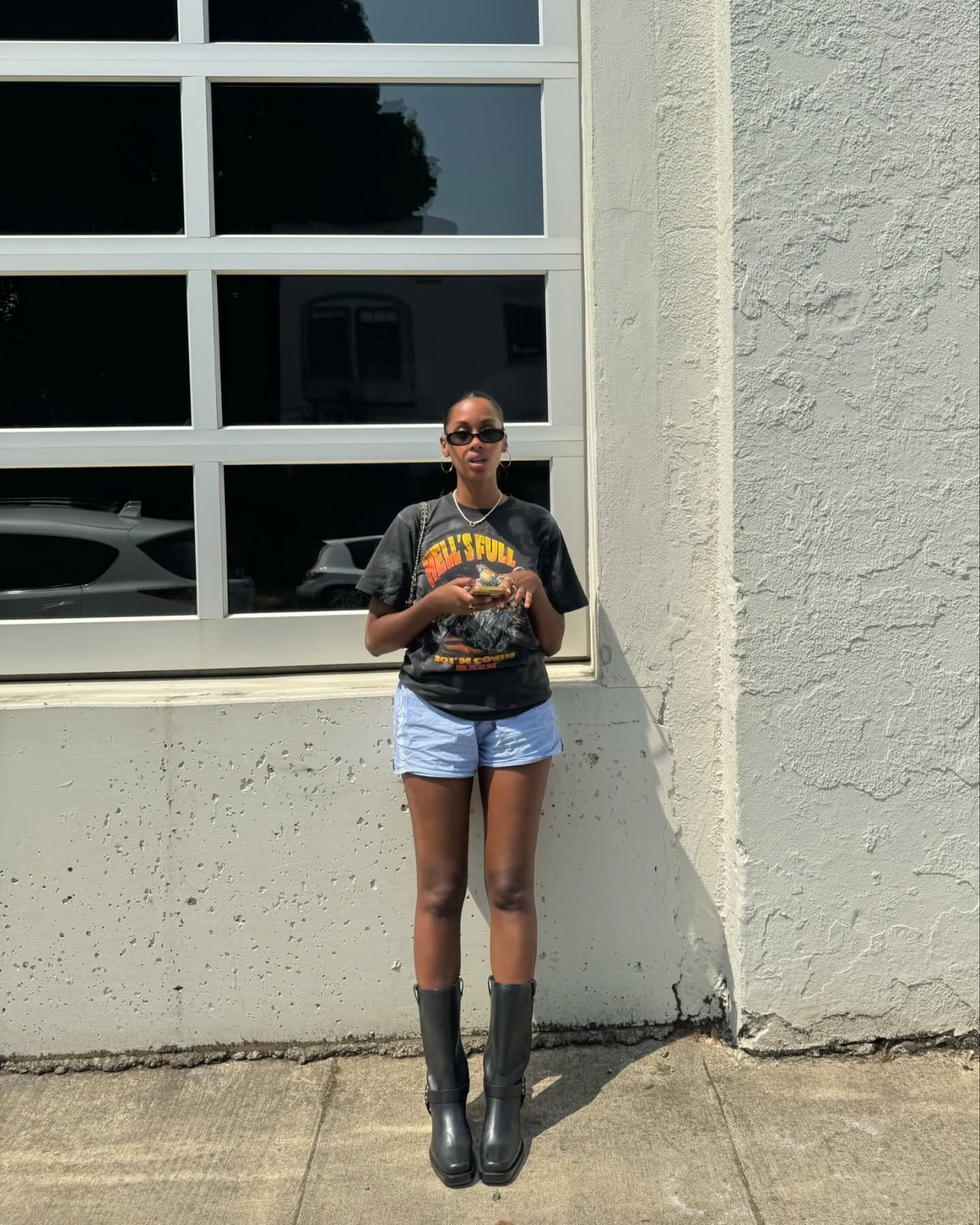 Pessoa em pé, usando camiseta preta e shorts azuis, e segurando celular. Ideal para looks contemporâneos.