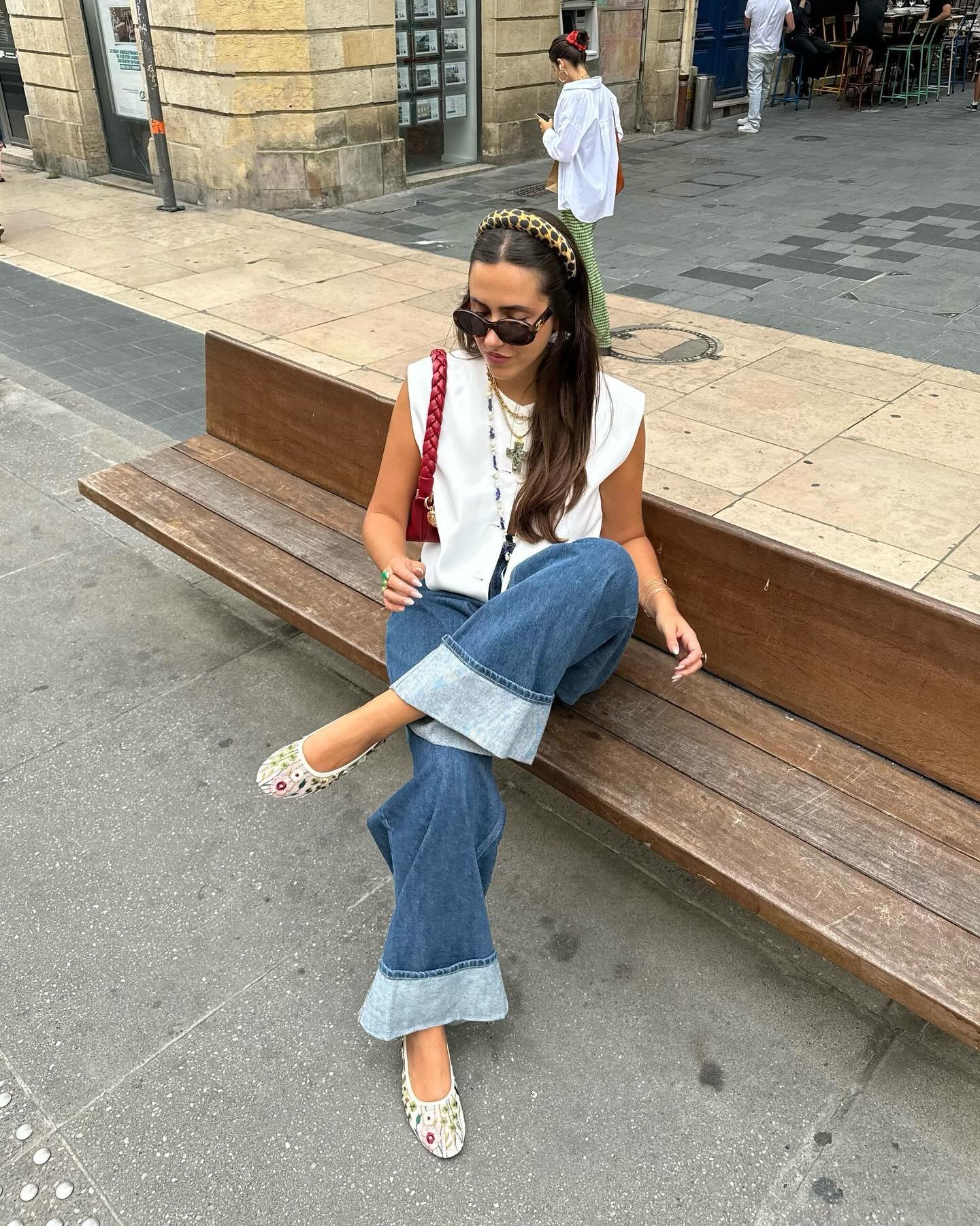Mulher sentada em um banco de pedra, usando jeans largos, blusa branca e acessórios elegantes.
