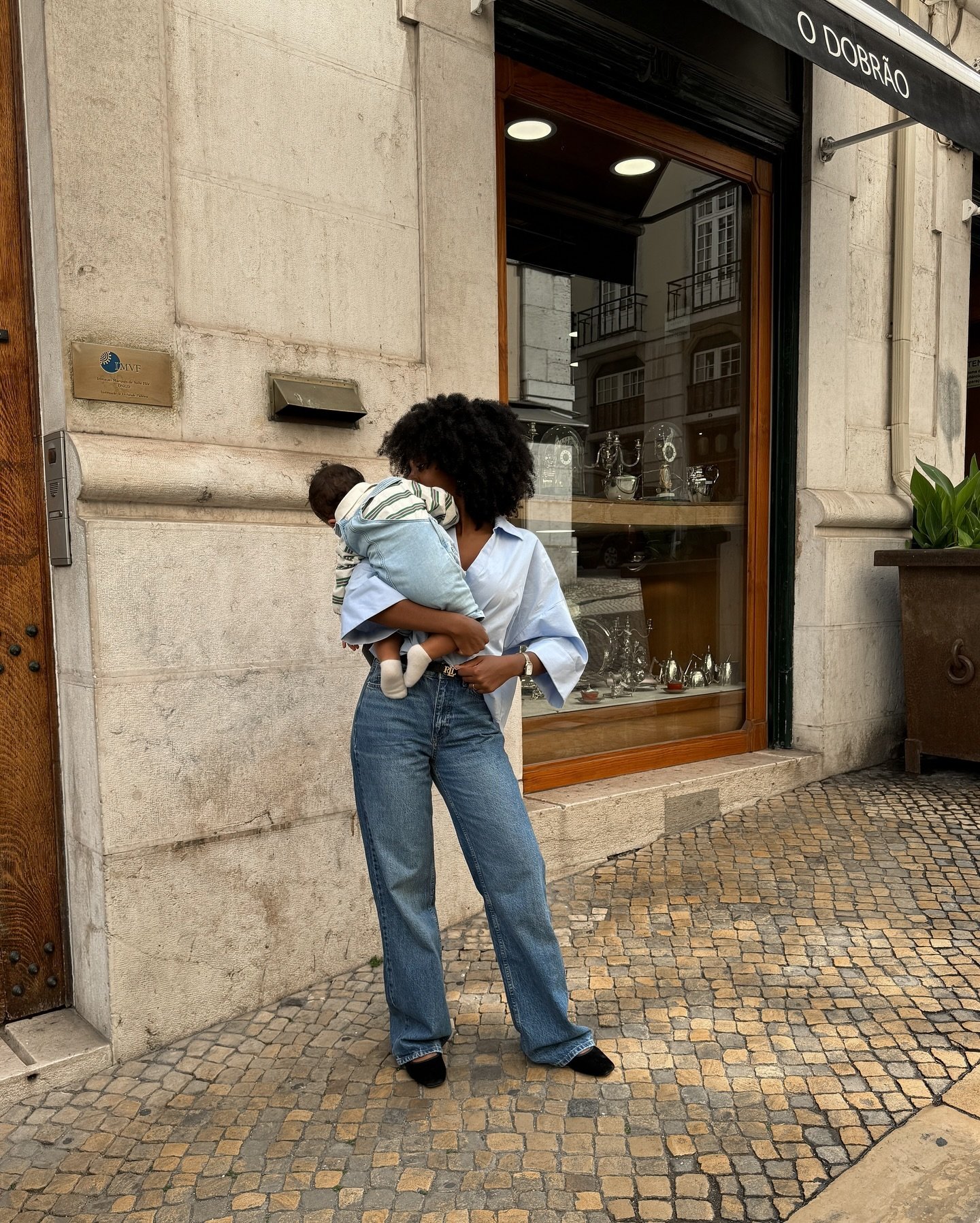 Mulher vestindo jeans e camisa azul segura um bebê na calçada, criando looks confortáveis para quem amamenta.