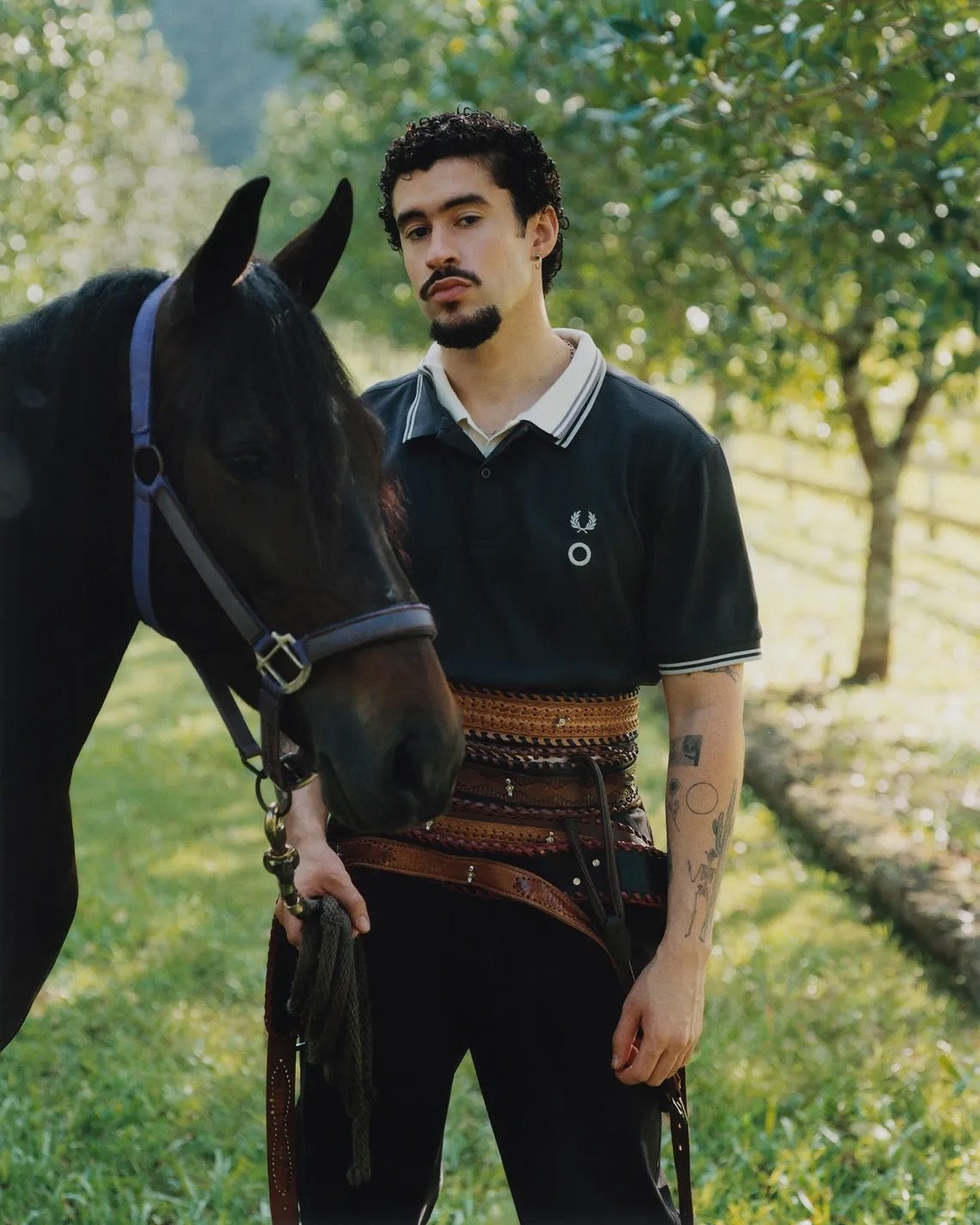 Jovem ao lado de um cavalo em cenário rural, veste polo preta e cinto largo, tendência esperada na moda de 2025.