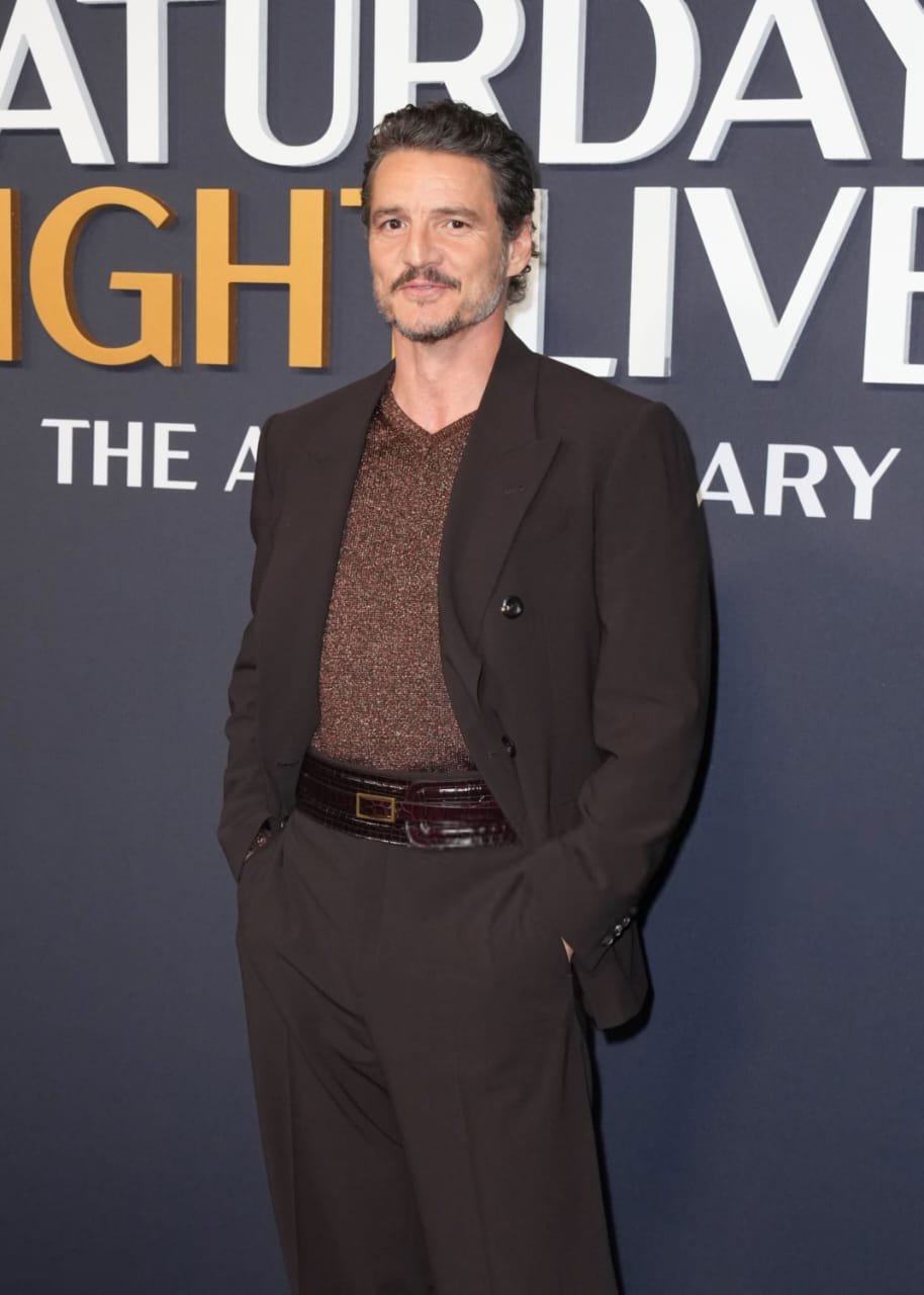 Homem vestindo terno marrom escuro e camisa cintilante, em frente a um painel de fundo com letras grandes.