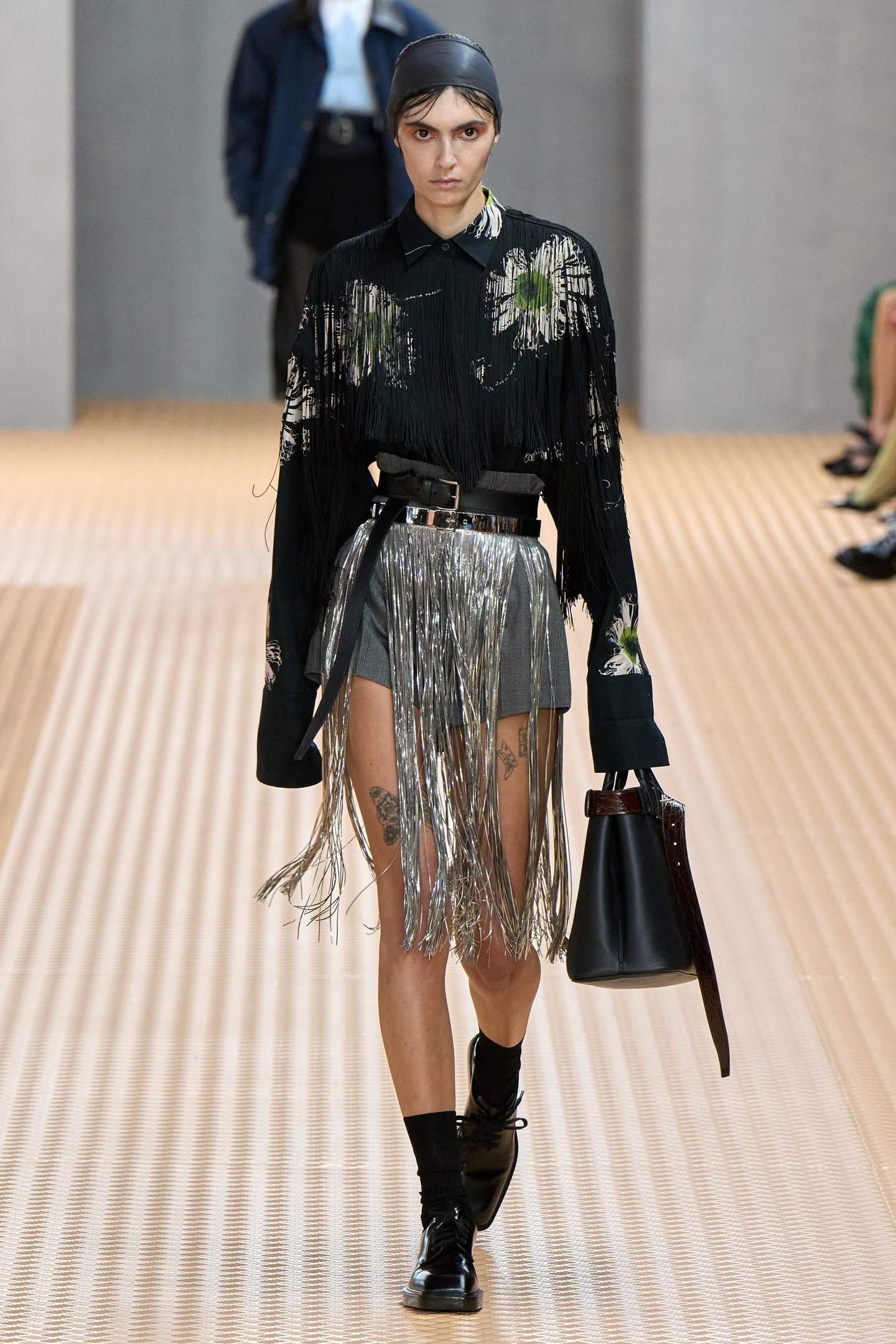 Modelo desfilando com roupas futuristas, incluindo franjas metálicas, em tendência de moda de 2025.