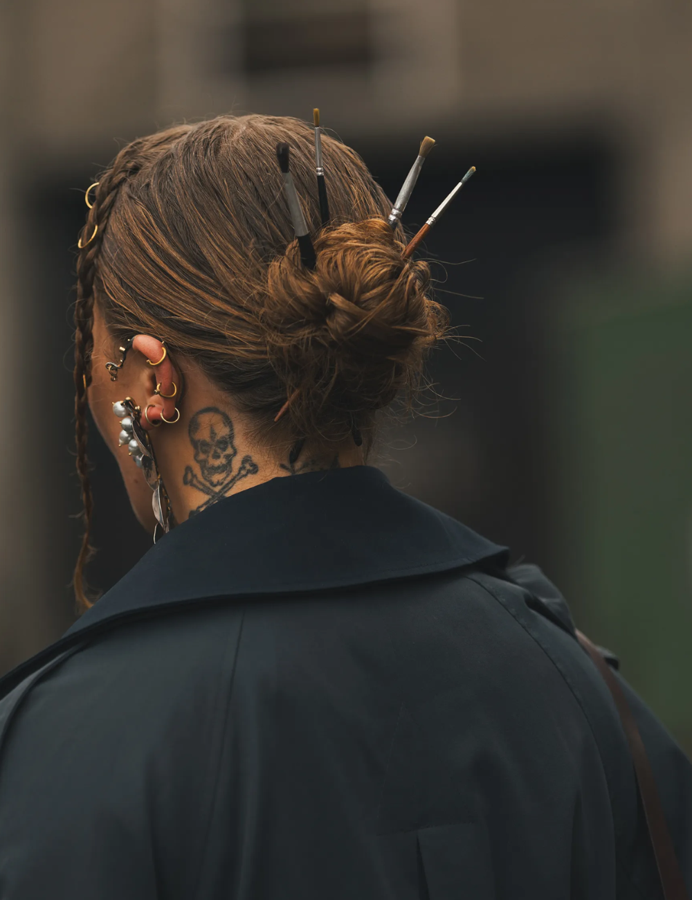 Penteado com pincéis inseridos, acessórios maximalistas no cabelo, e tatuagem de caveira no pescoço.