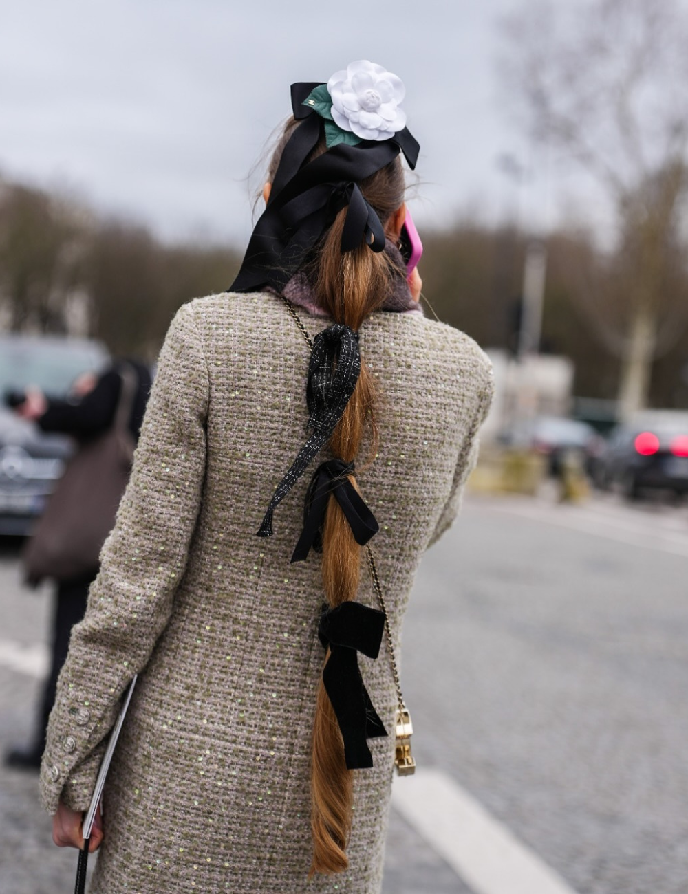 Penteado com trança decorada por laços pretos e flor branca, exemplo de acessórios maximalistas no cabelo.