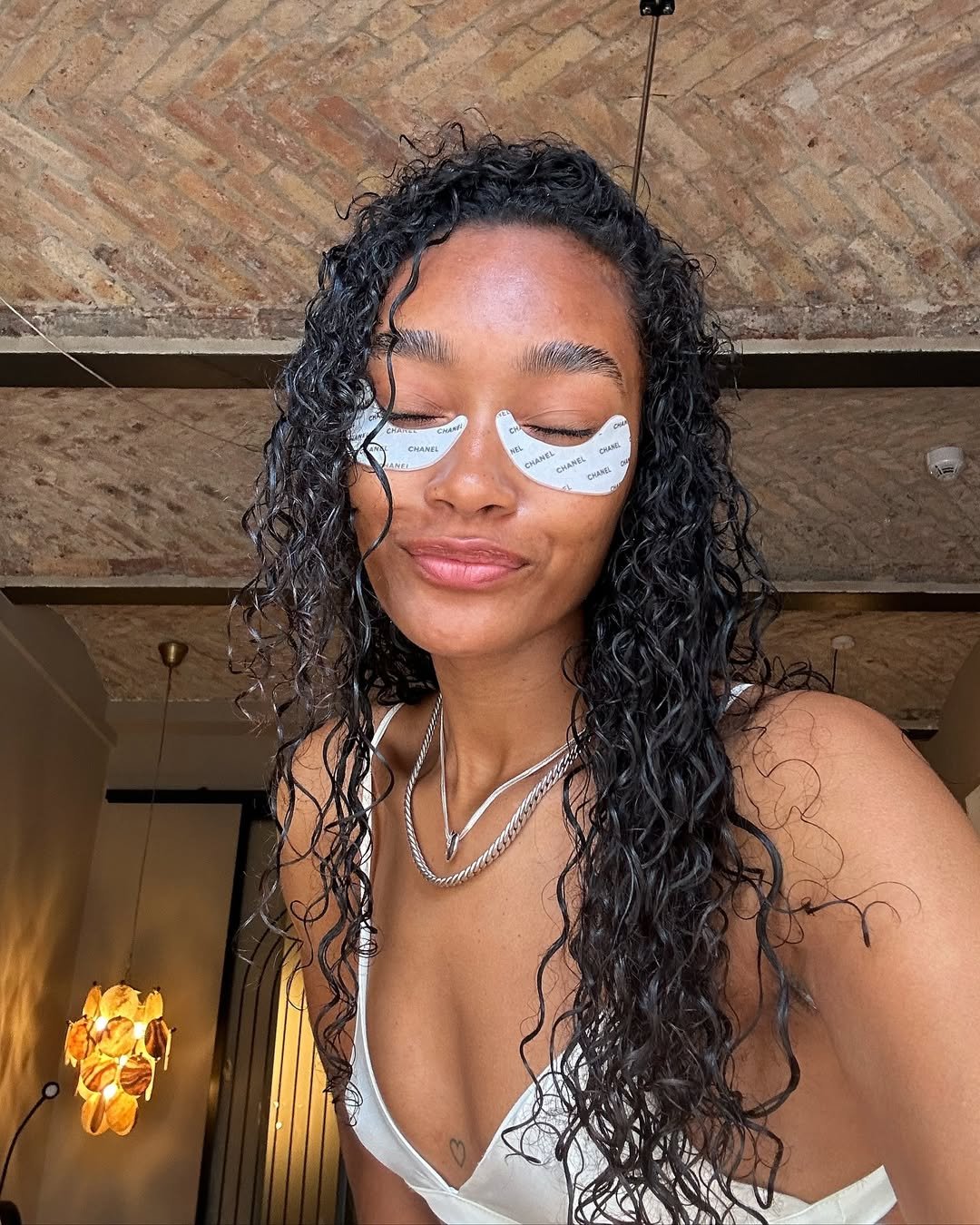 AHA (alfa-hidroxiácidos)  Mulher com curvatura nos cabelos usa adesivos faciais sob os olhos, sorrindo levemente em ambiente interno.