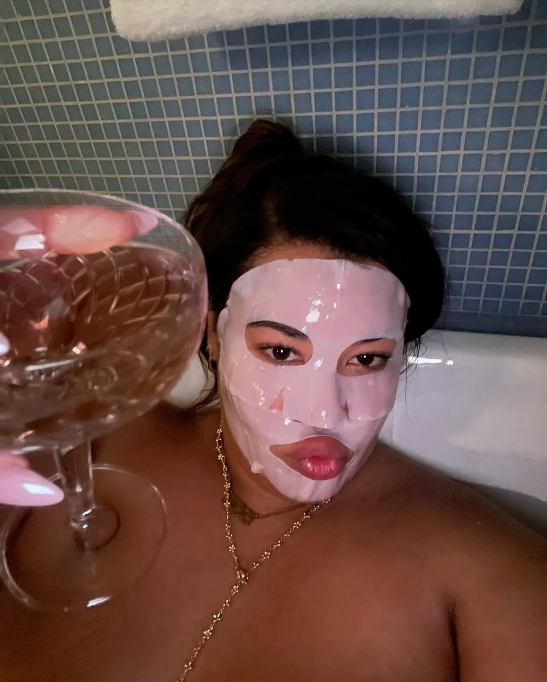 Pessoa relaxando em banheira, usando máscara facial e segurando taça. Momento de autocuidado e relaxamento. AHA (alfa-hidroxiácidos)
