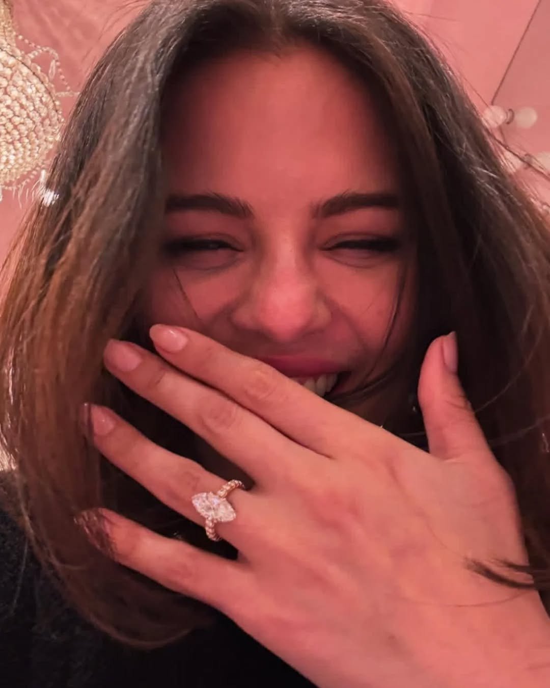 Mulher sorrindo cobre a boca, exibindo anel brilhante no dedo anular.