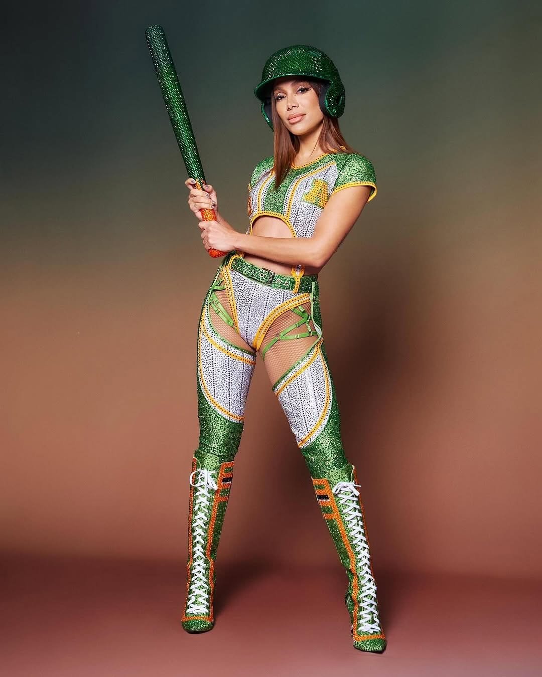 Anitta em traje temático de beisebol, com taco e capacete verdes brilhantes.