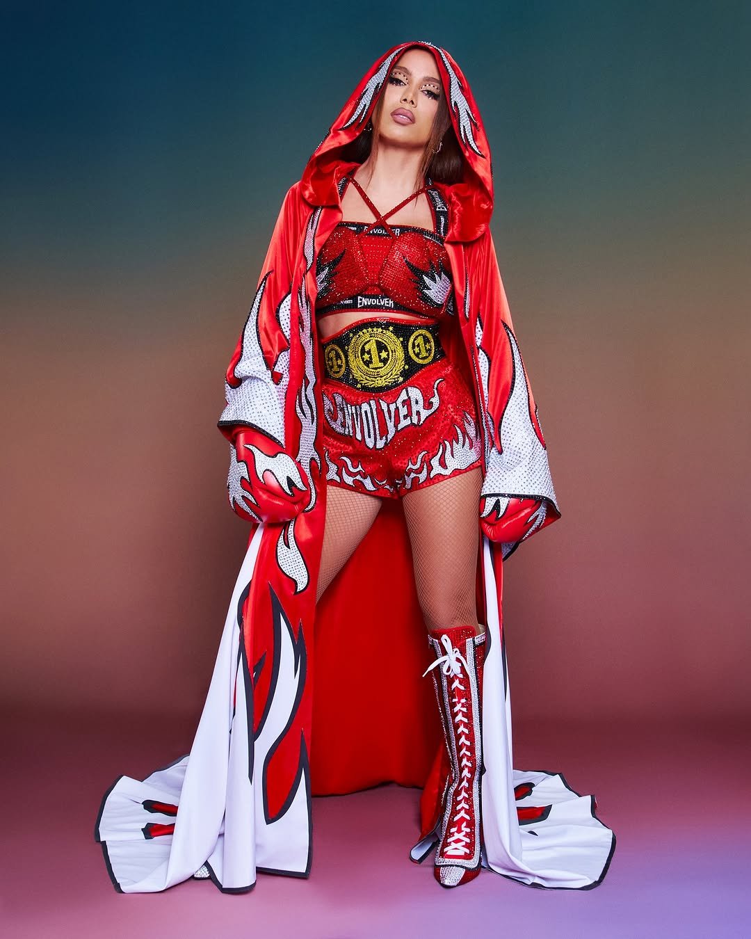 Anitta com traje vermelho e capa estampada, em pé, em estilo vibrante e ousado com tema de chamas.