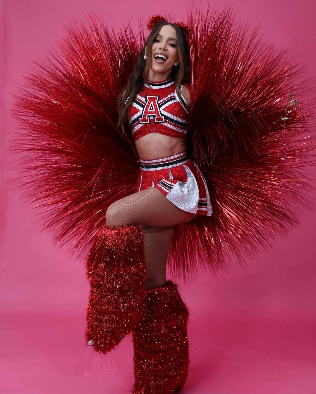 Anitta com roupa de animadora de torcida vermelha brilhante, posando em fundo rosa.