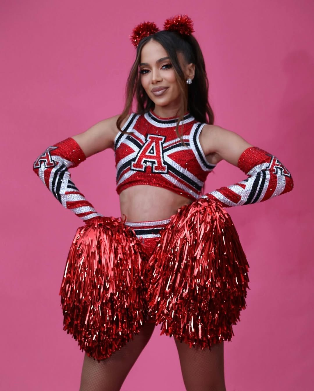Anitta com pompons vermelhos exibe uniforme vibrante em fundo rosa.