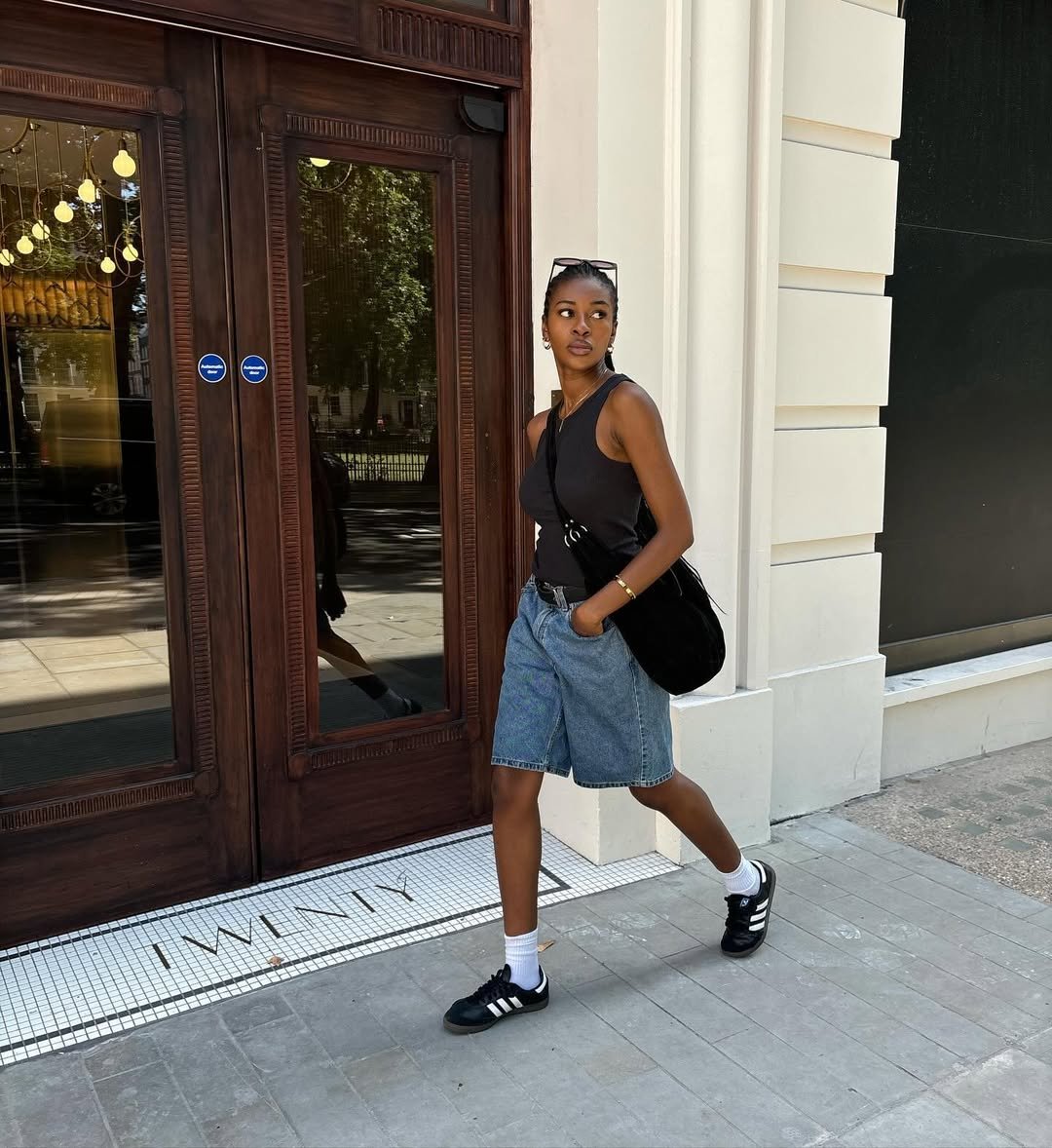 Pessoa andando na calçada, usando regata preta e bermuda jeans, com tênis preto e bolsa a tiracolo.