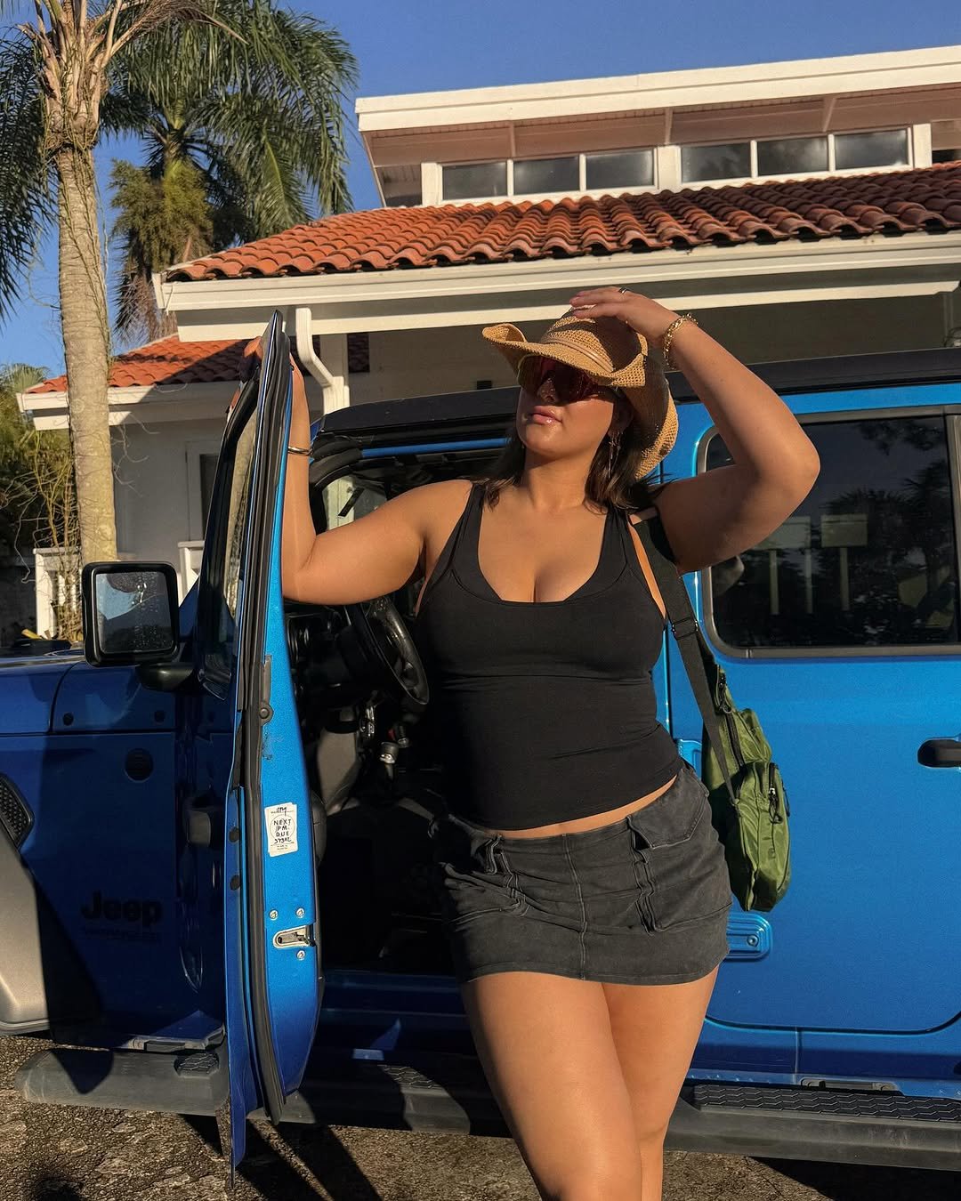 Pessoa usando blusa preta e chapéu de palha, apoiada em um carro azul, com uma casa ao fundo.