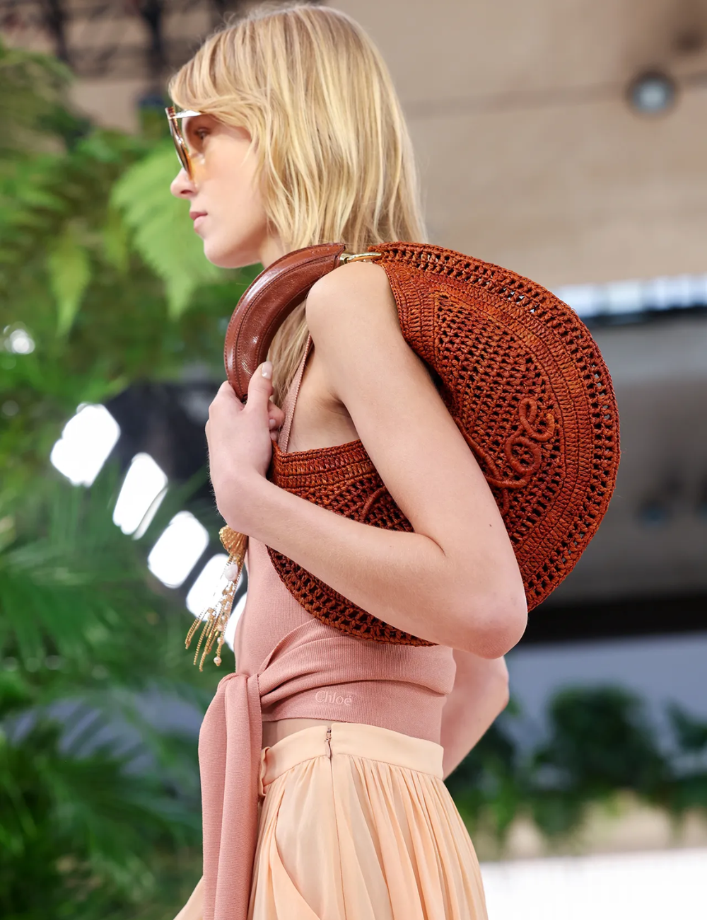 Mulher com bolsa de crochê marrom no ombro, vestindo roupa elegante em desfile, referência de bolsas tendência 2025.