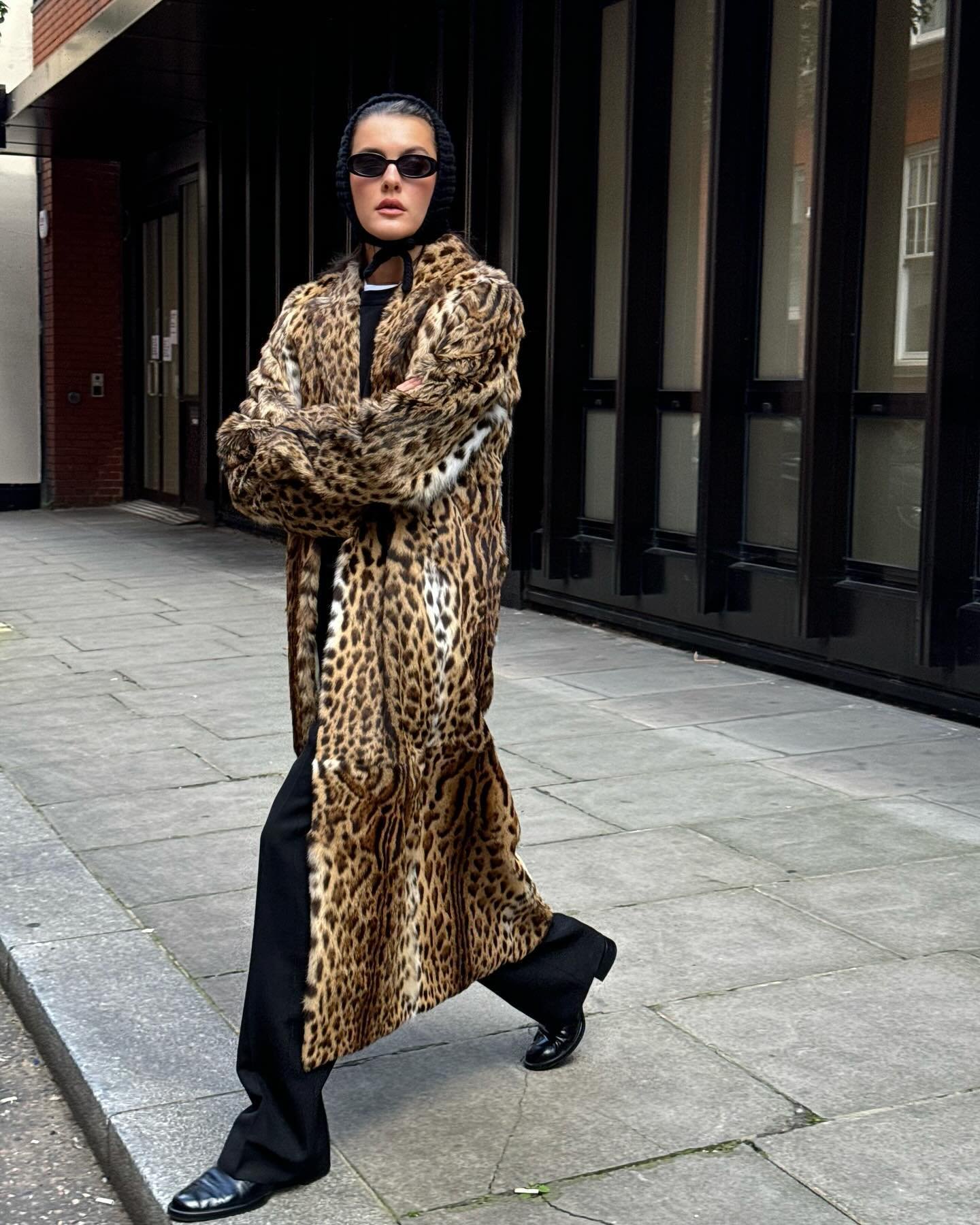 Pessoa usa casaco com estampa de leopardo, óculos escuros e bonnet preto em rua de cidade.