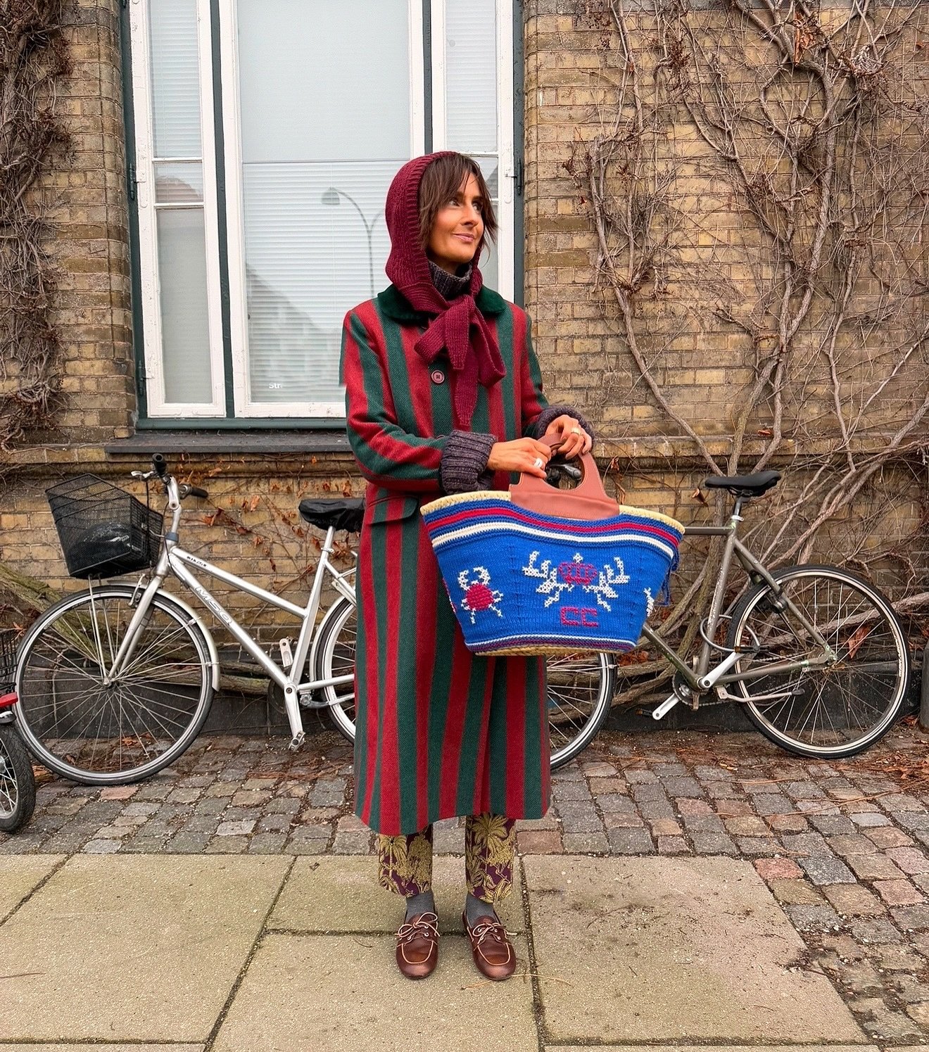 Mulher com casaco listrado e cachecol estilo bonnet, segurando uma bolsa colorida ao lado de bicicletas.