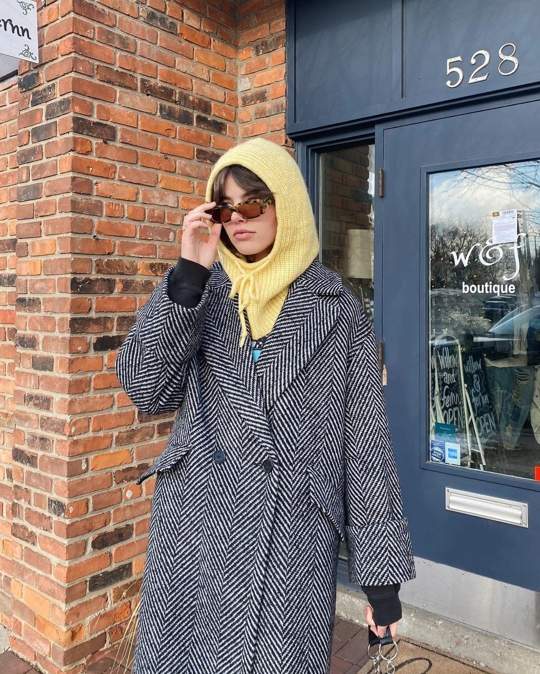 Pessoa com casaco estampado e capuz amarelo estilo bonnet, em frente a uma loja com fachada de tijolos.