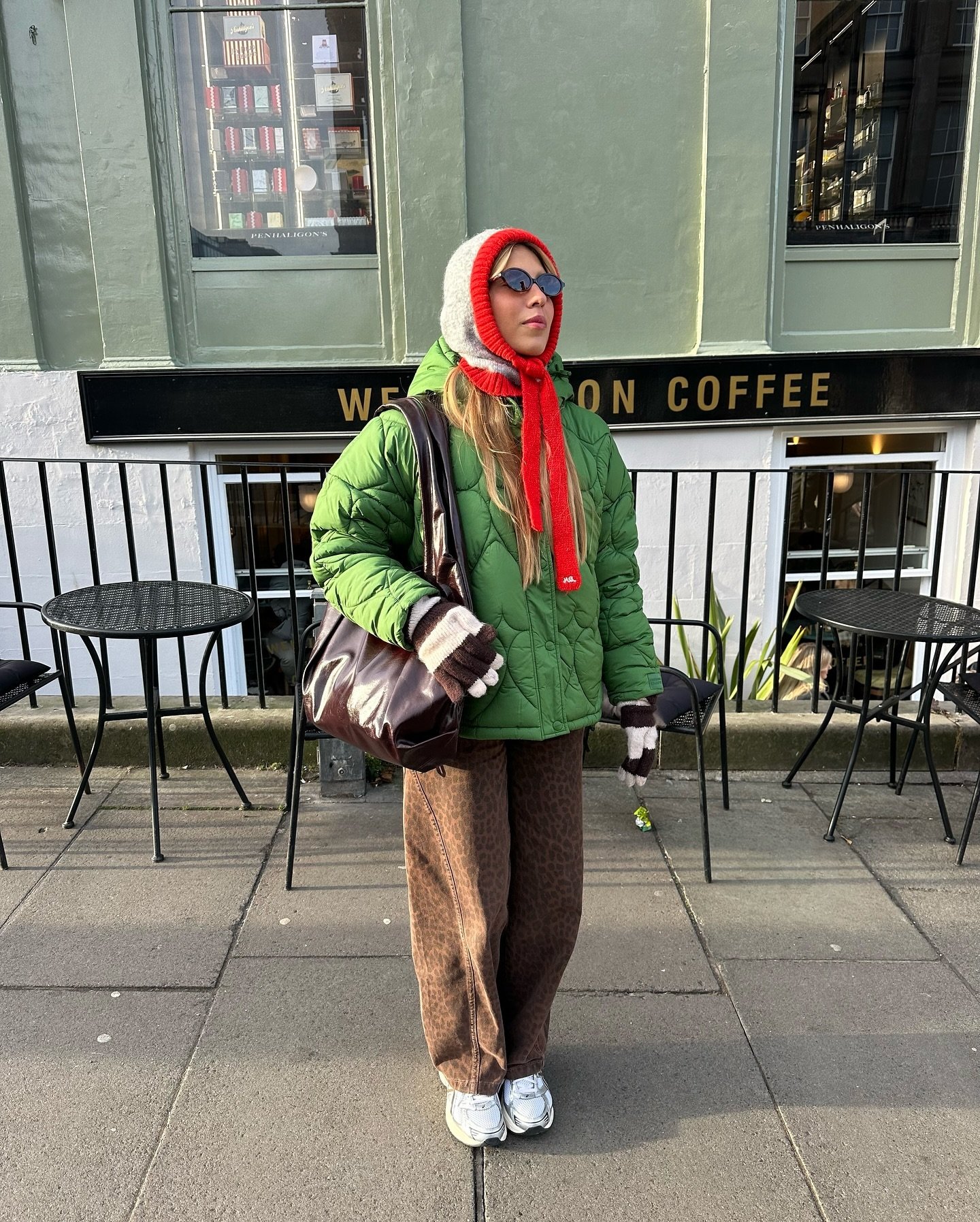 Mulher com casaco verde e chapéu de inverno vermelho e cinza, em frente a um café.