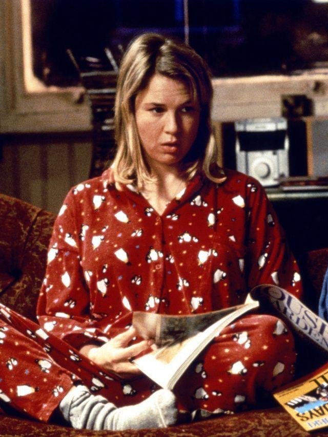 Mulher de pijama vermelho com estampa sentada no sofá, segurando revistas. Ambiente lembra cenas de Bridget Jones.