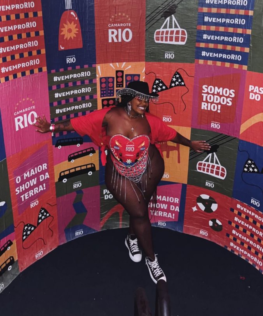 Pessoa sorrindo em um camarote decorado com cartazes coloridos do Rio. Roupas vibrantes e adereços carnavalescos. previsões de carnaval
