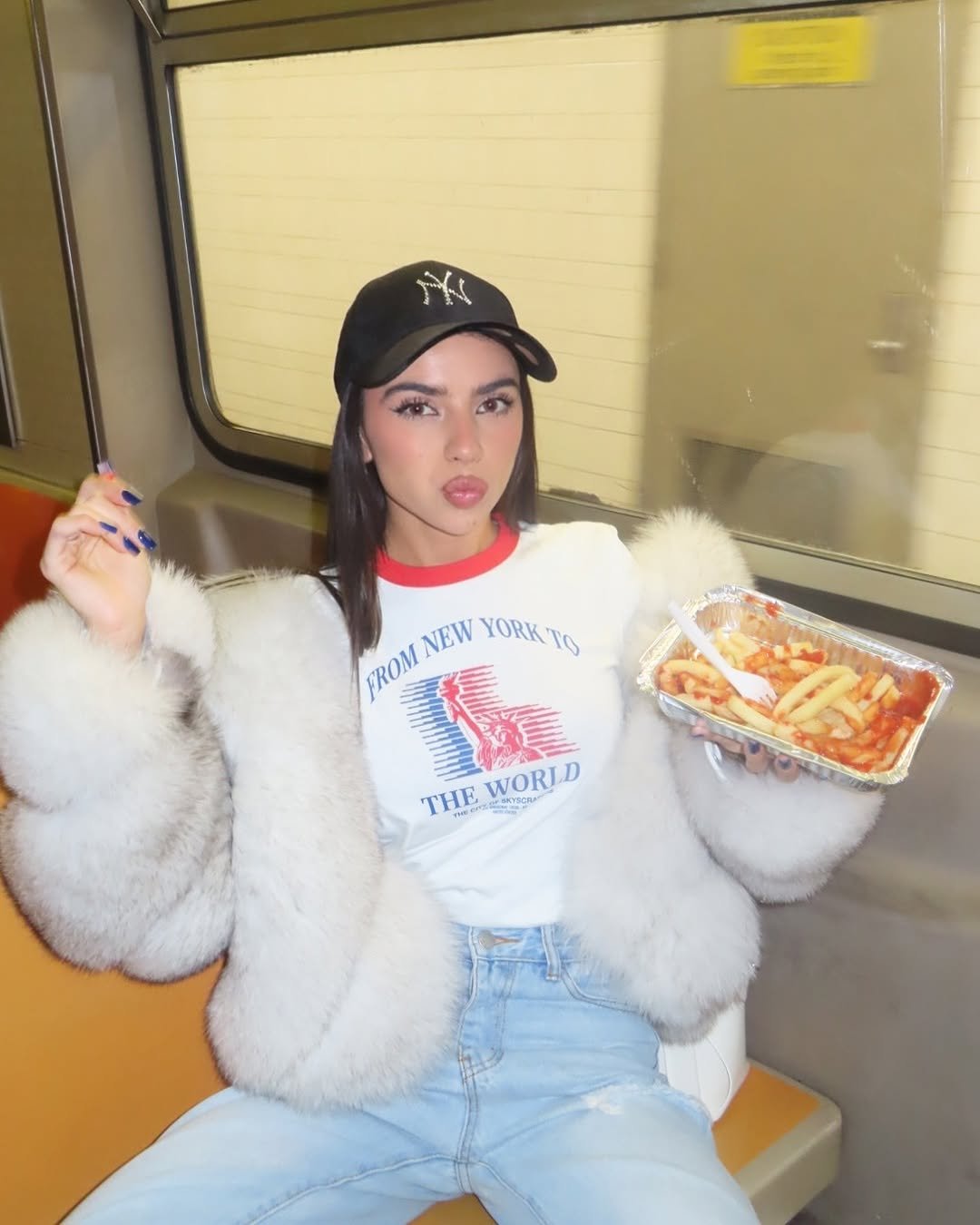 Mulher em metrô, vestindo casaco de pele, camiseta e boné, segura comida. Visual como um combo de estilo.