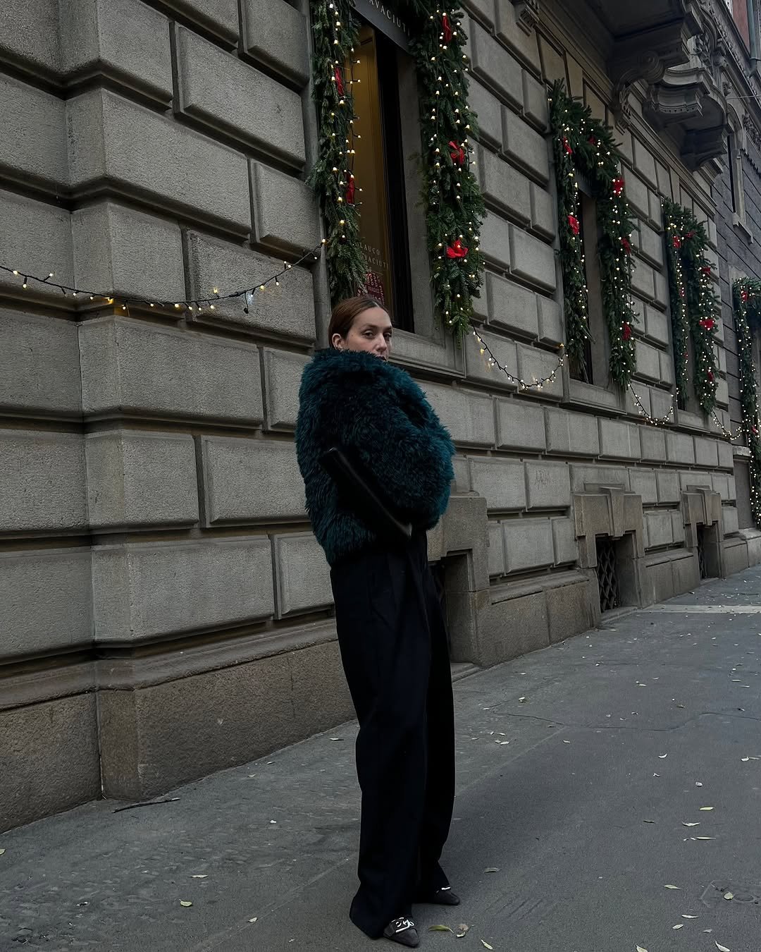 Pessoa com casaco peludo azul escuro em frente a prédio decorado com guirlandas natalinas.