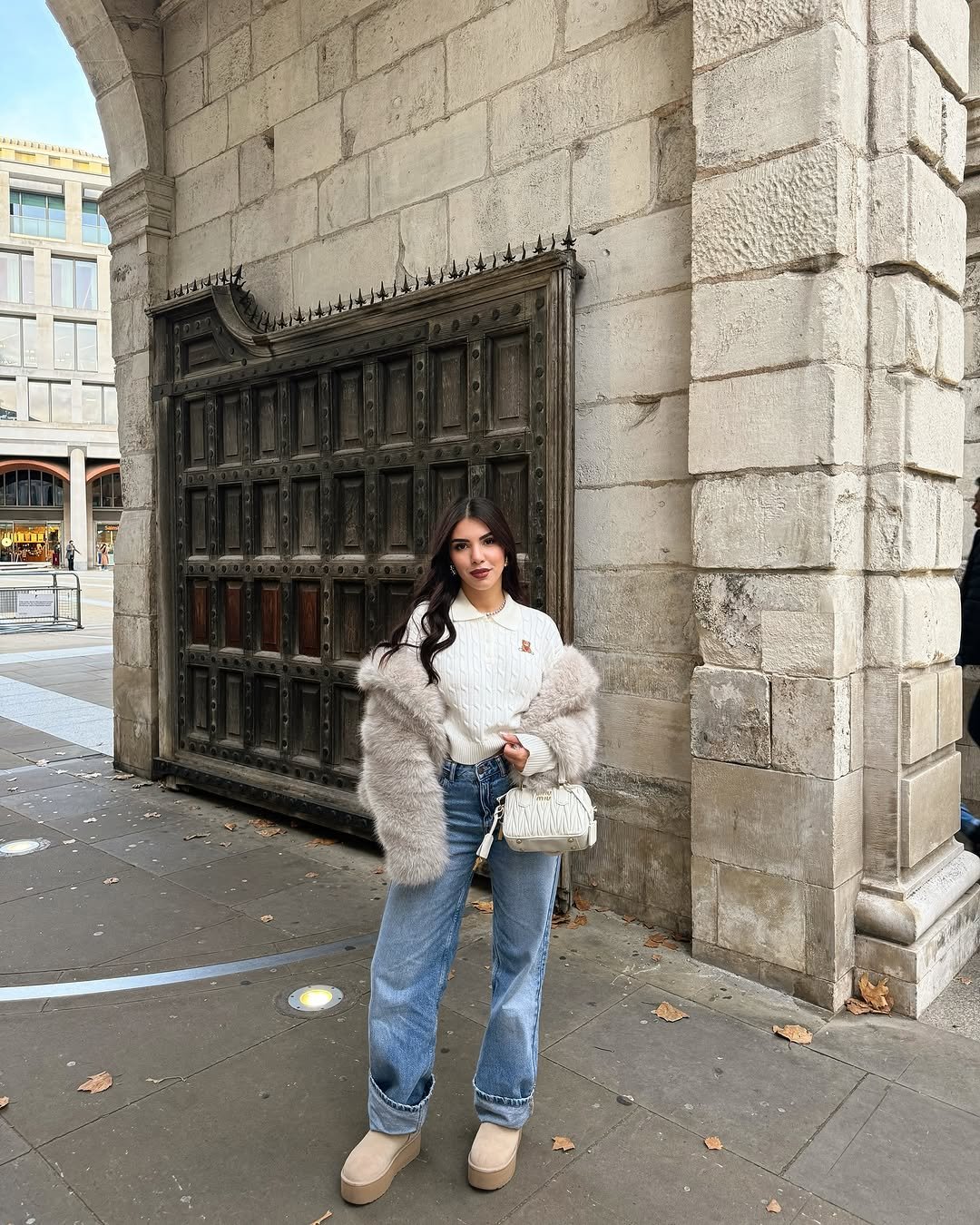 Mulher posando com roupa elegante e casual em frente a uma porta antiga. O look combina jeans, malha e casaco felpudo. Combo de estilo