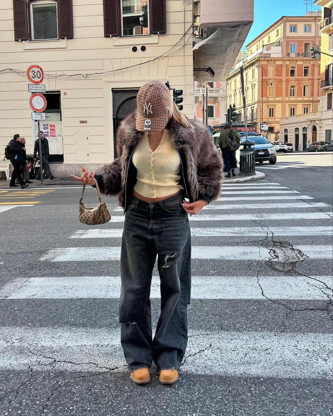 Pessoa cruzando a rua com um casaco peludo, blusa amarela e boné xadrez. Roupa forma um combo de estilo urbano.