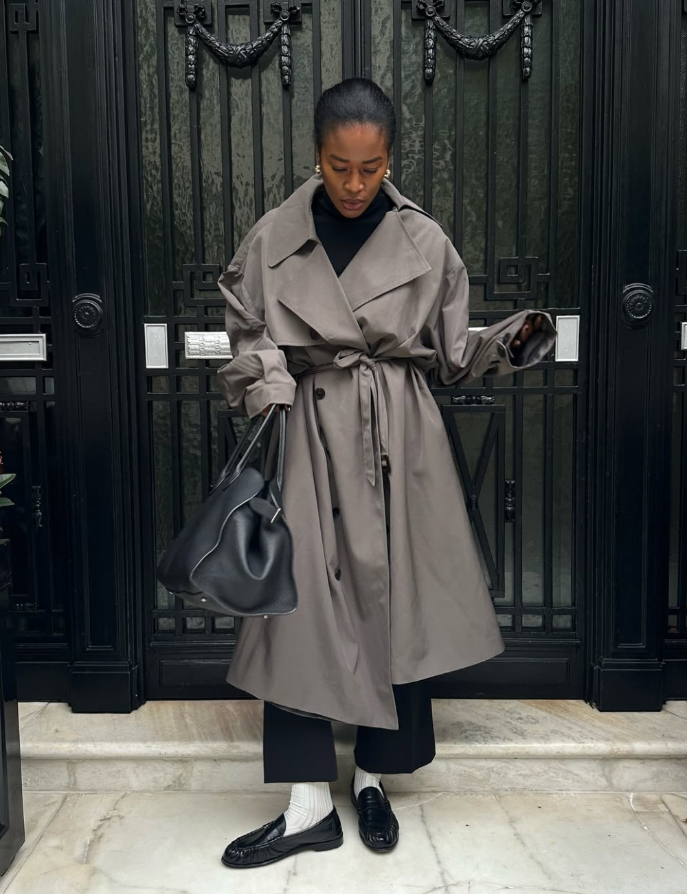 Pessoa usando trench coat cinza e carregando bolsa preta, com sapato elegante e curinga.