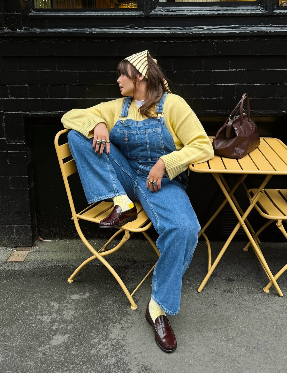 Pessoa sentada em cadeira amarela, vestindo macacão jeans, suéter amarelo e sapato vinho sofisticado. Bolsa ao lado.