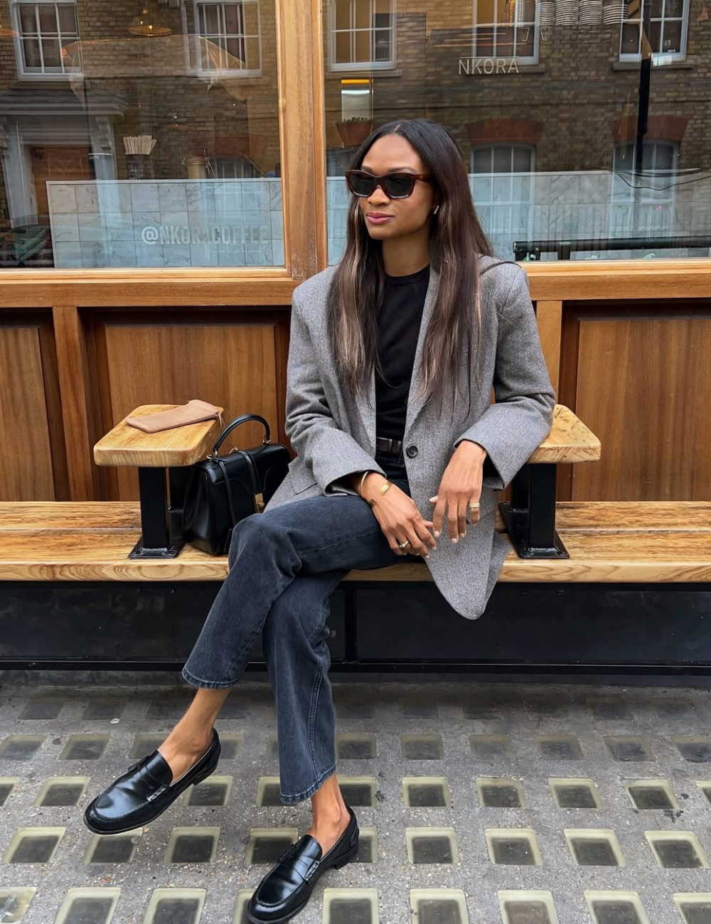 Pessoa elegante sentada em banco de madeira, usando blazer cinza, calça preta e mocassins pretos. Sapato elegante e curinga.