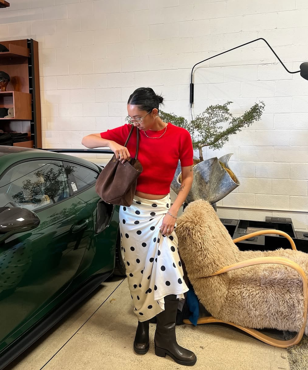 Mulher com saia estampa poá, blusa vermelha e botas pretas ao lado de carro verde e cadeira bege.
