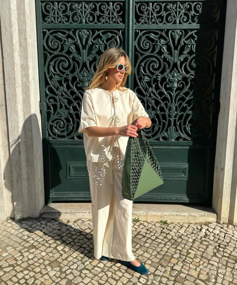 Mulher com óculos de sol e bolsa verde, usando sapatilhas, em frente a um portão decorativo. Dicas de como usar sapatilhas em 2025.