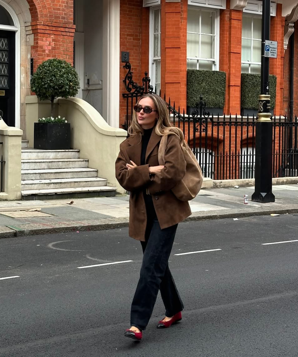 Mulher andando em rua urbana, usa casaco marrom, jeans e sapatilhas vermelhas, combinação elegante para 2025.