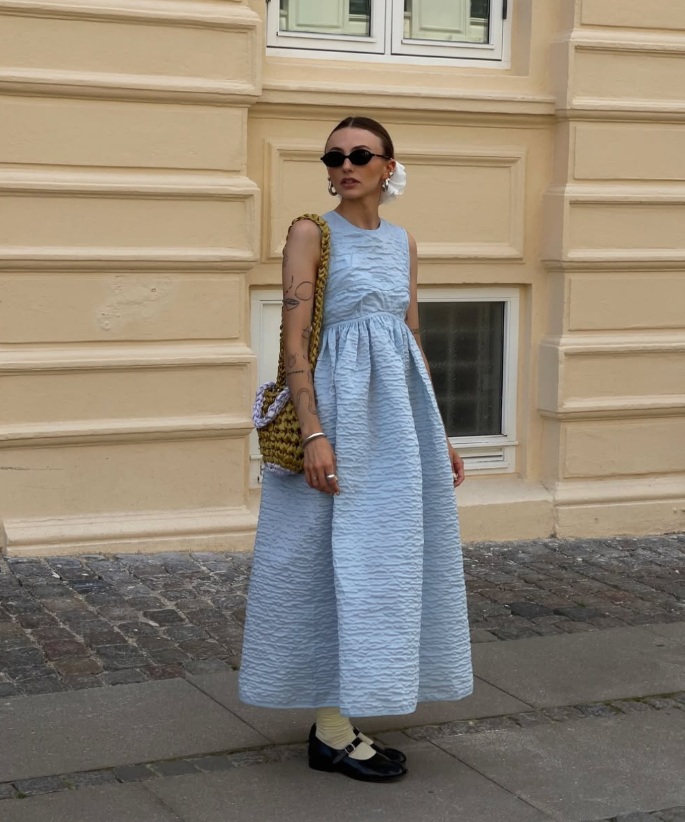 Mulher com vestido azul texturizado e sapatilhas estilo vintage. Ela está em frente a um edifício bege.