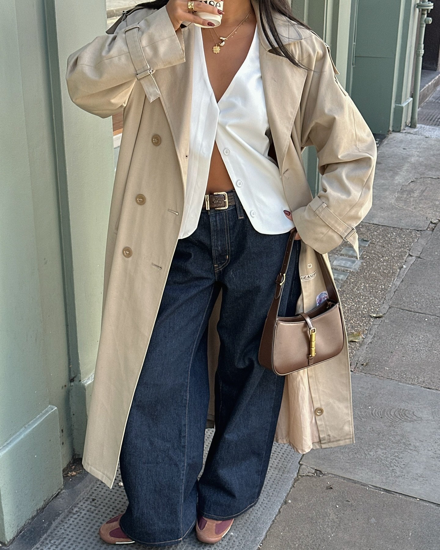 Pessoa usa casaco bege, camisa branca, calça jeans, bolsa marrom e tênis marrons na rua.