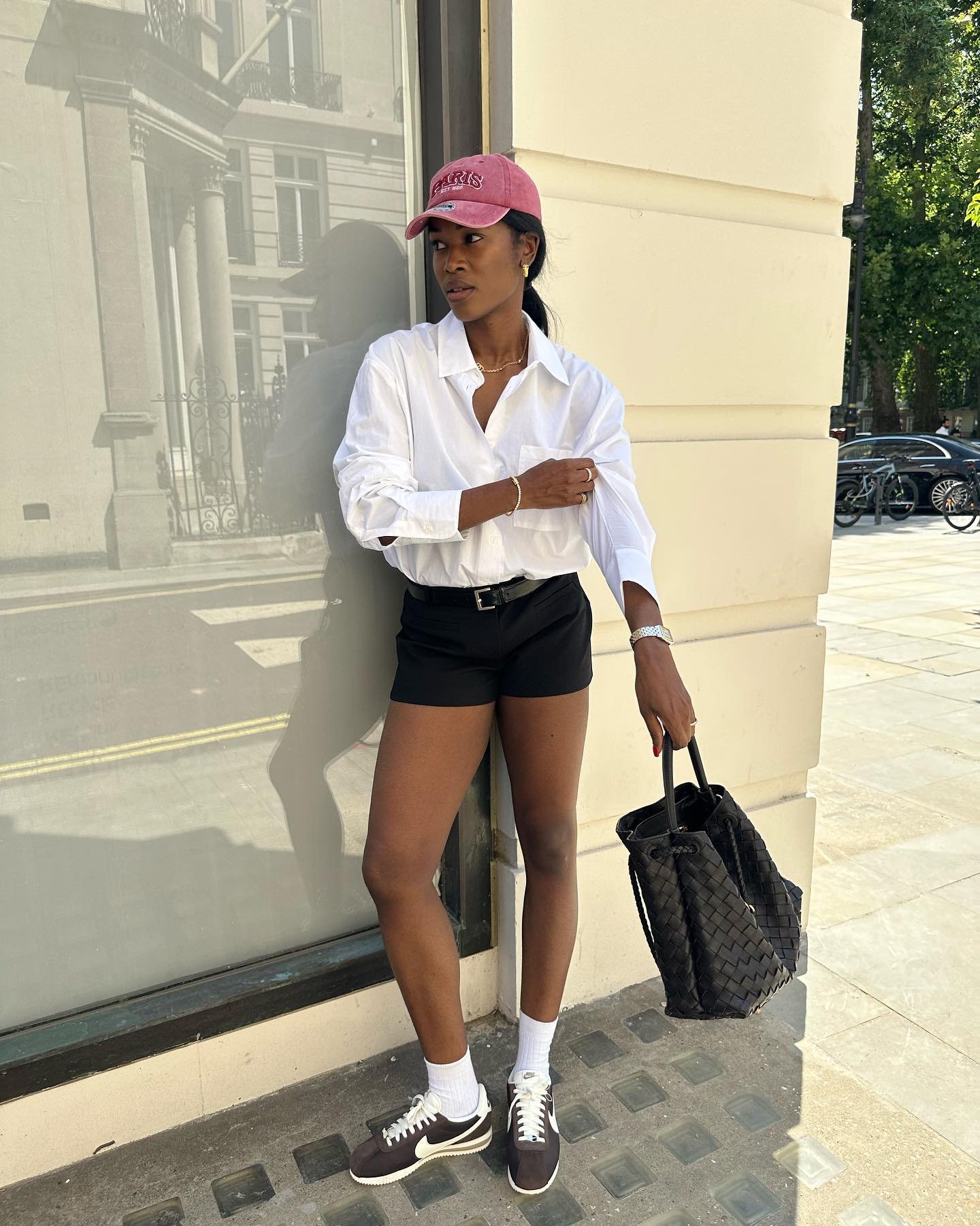 Pessoa com boné rosa, camisa branca e shorts pretos encostada em uma parede. Usa tênis marrom e carrega bolsa preta.