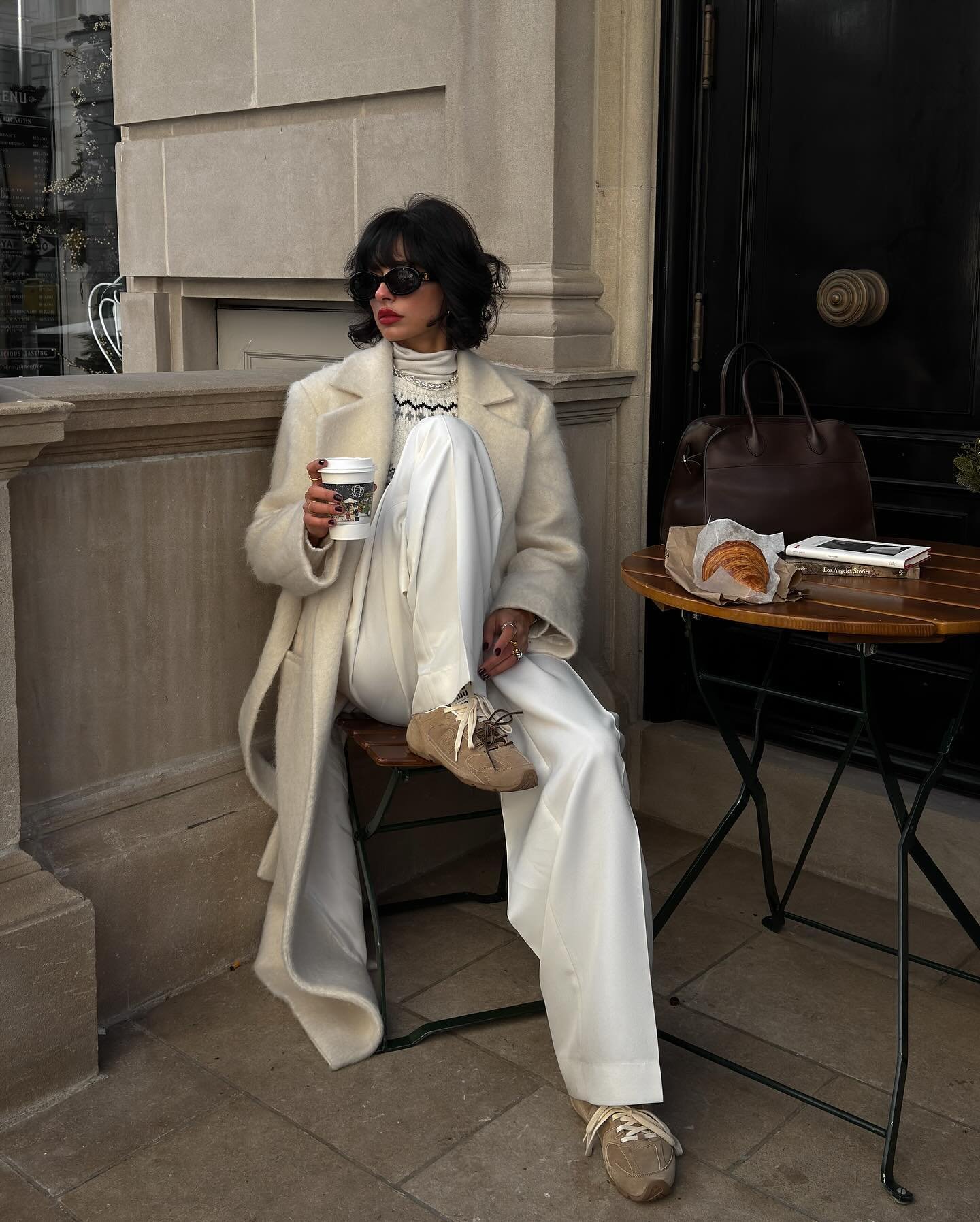 Pessoa elegante em casaco claro e tênis marrom, sentada, segurando café em frente a mesa com croissant.