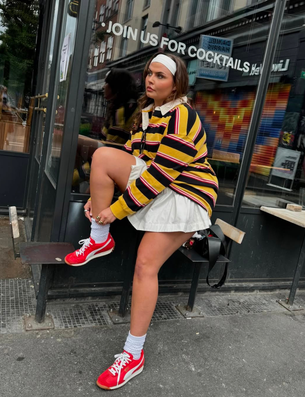 Pessoa sentada em frente a uma loja, usando blusa listrada e tênis vermelhos, destacando cores de tênis.