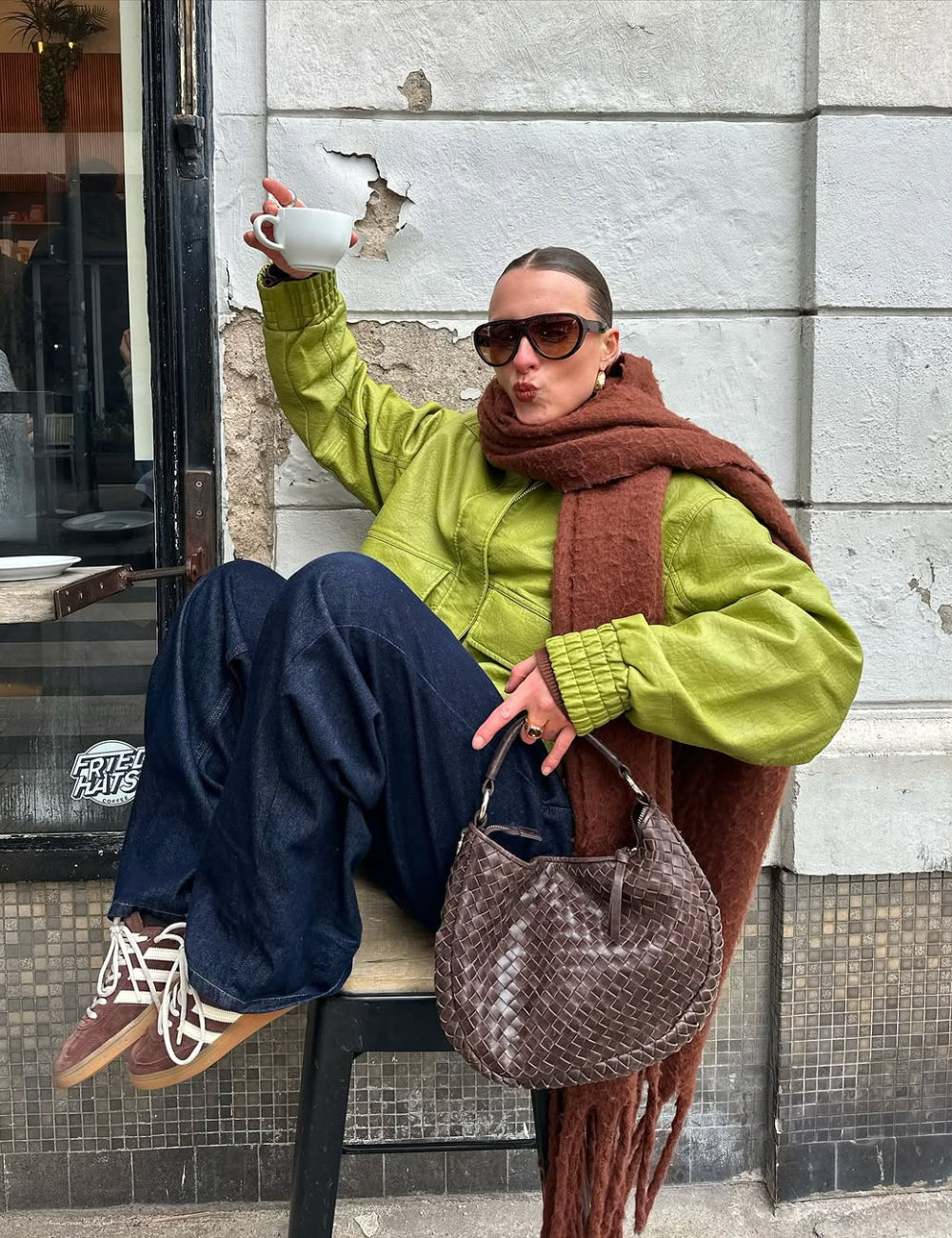 Pessoa sentada, usando jaqueta verde, cachecol marrom, calças jeans escuras. Tênis marrons com detalhes brancos.